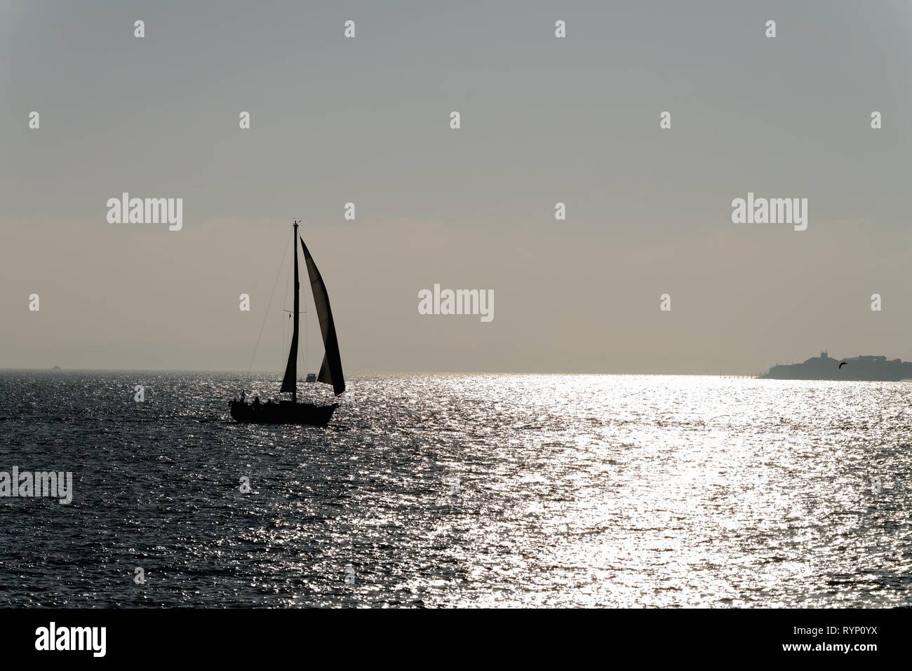 Silhouette einer Yacht segeln auf den Solent in Portsmouth Hampshire, gegen die Reflexion von Sonnenlicht auf dem Meer, England Großbritannien Stockfoto
