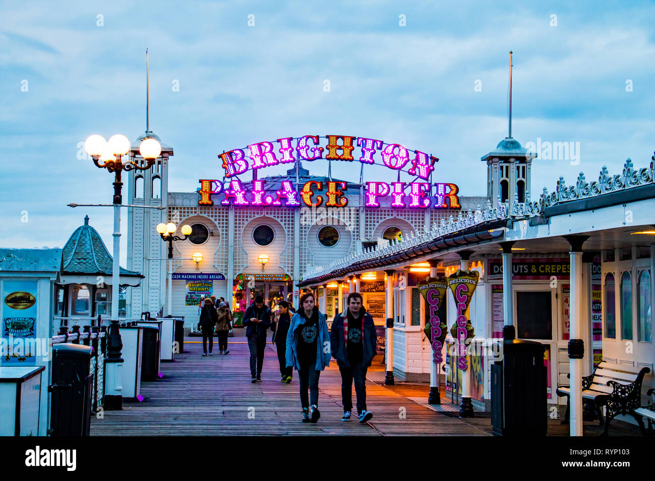 Menschen flanieren auf Brighton Palace Pier am Abend mit seinen bunten Zeichen in der gedimmtem Licht glühenden Stockfoto
