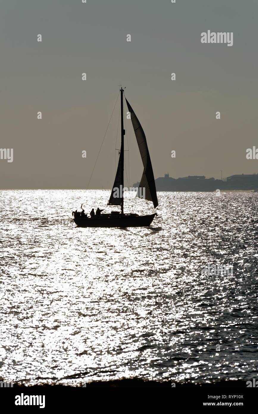 Silhouette einer Yacht segeln auf den Solent in Portsmouth Hampshire, gegen die Reflexion von Sonnenlicht auf dem Meer, England Großbritannien Stockfoto