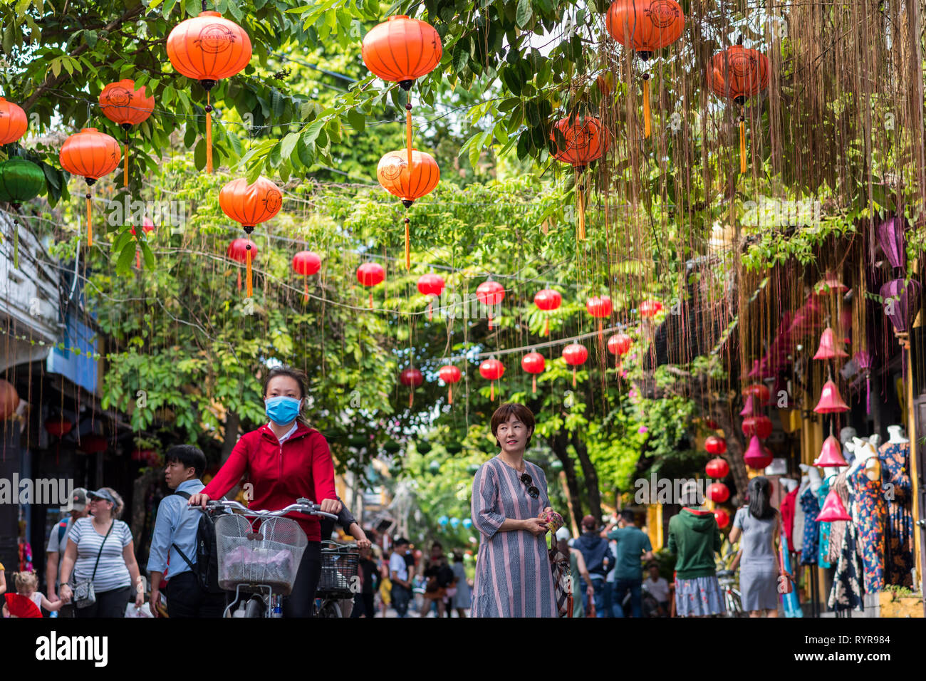 Hoi An, Vietnam - Oktober 23, 2018: eine Straße mit Perlen von Laternen hängen von Vordach von üppigem Laub und eine reife Frau in Kleid & ein Mädchen auf dem Fahrrad. Stockfoto