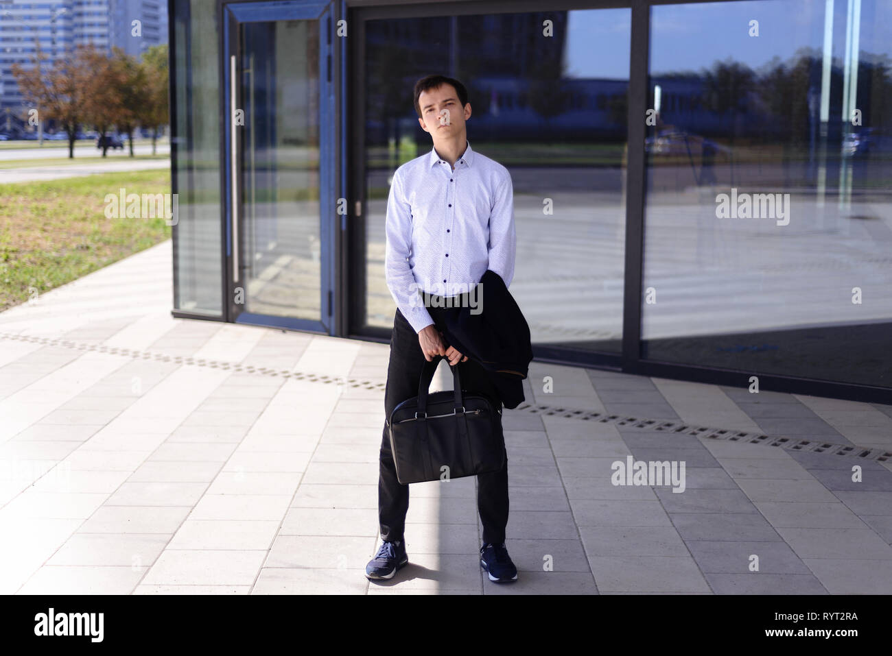 Junge Unternehmer Kerl steht in der Nähe von Gebäude mit großen Fenstern. Mann im Hemd hält einen Aktenkoffer und Jacke in den Händen Stockfoto