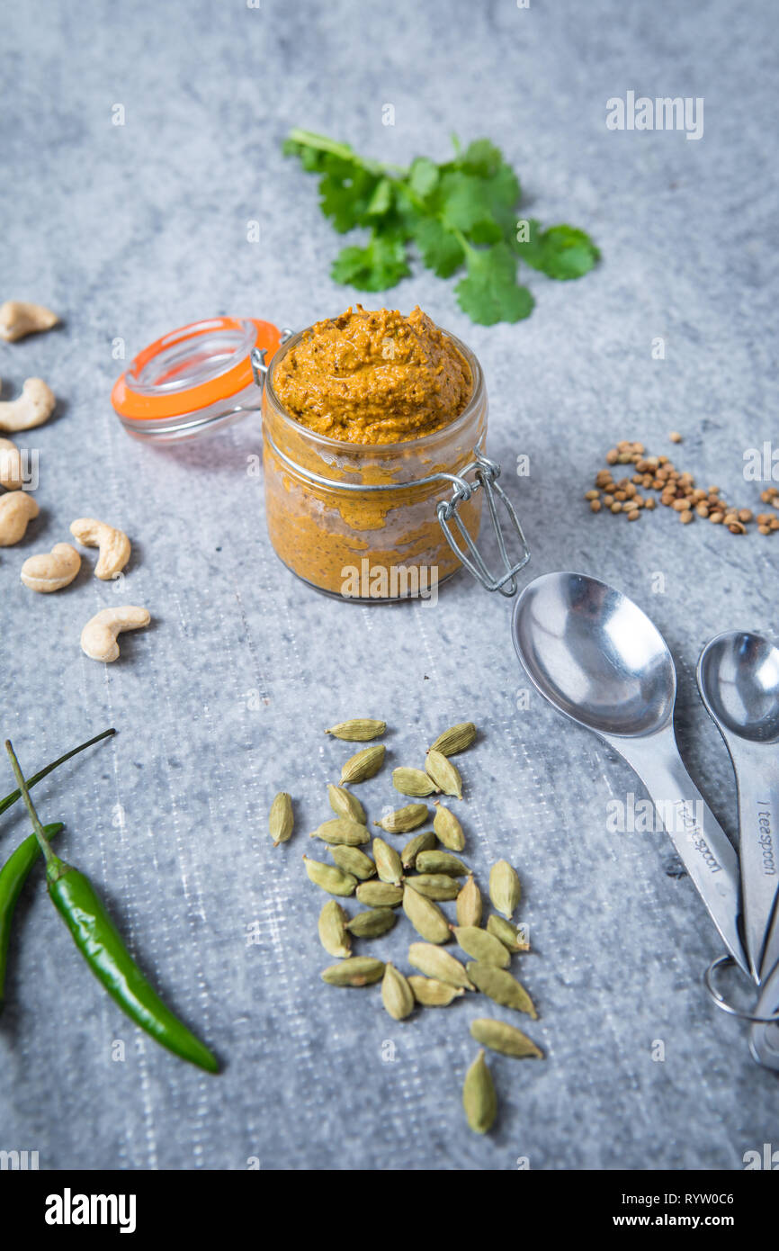 Curry Paste in ein Marmeladenglas mit Zutaten für currypaste auf grauem Hintergrund. Curry Paste, grüner Pfeffer, Kardamom, Cashewnüsse, Löffel, Koriander Stockfoto