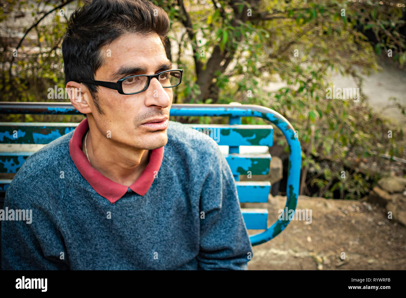 Outdoor Bild von asiatischen, indischen serene Bebrillte junger erwachsener Mann sitzt auf einer Parkbank und denken mit leeren Ausdruck während weg in Tag suchen Stockfoto