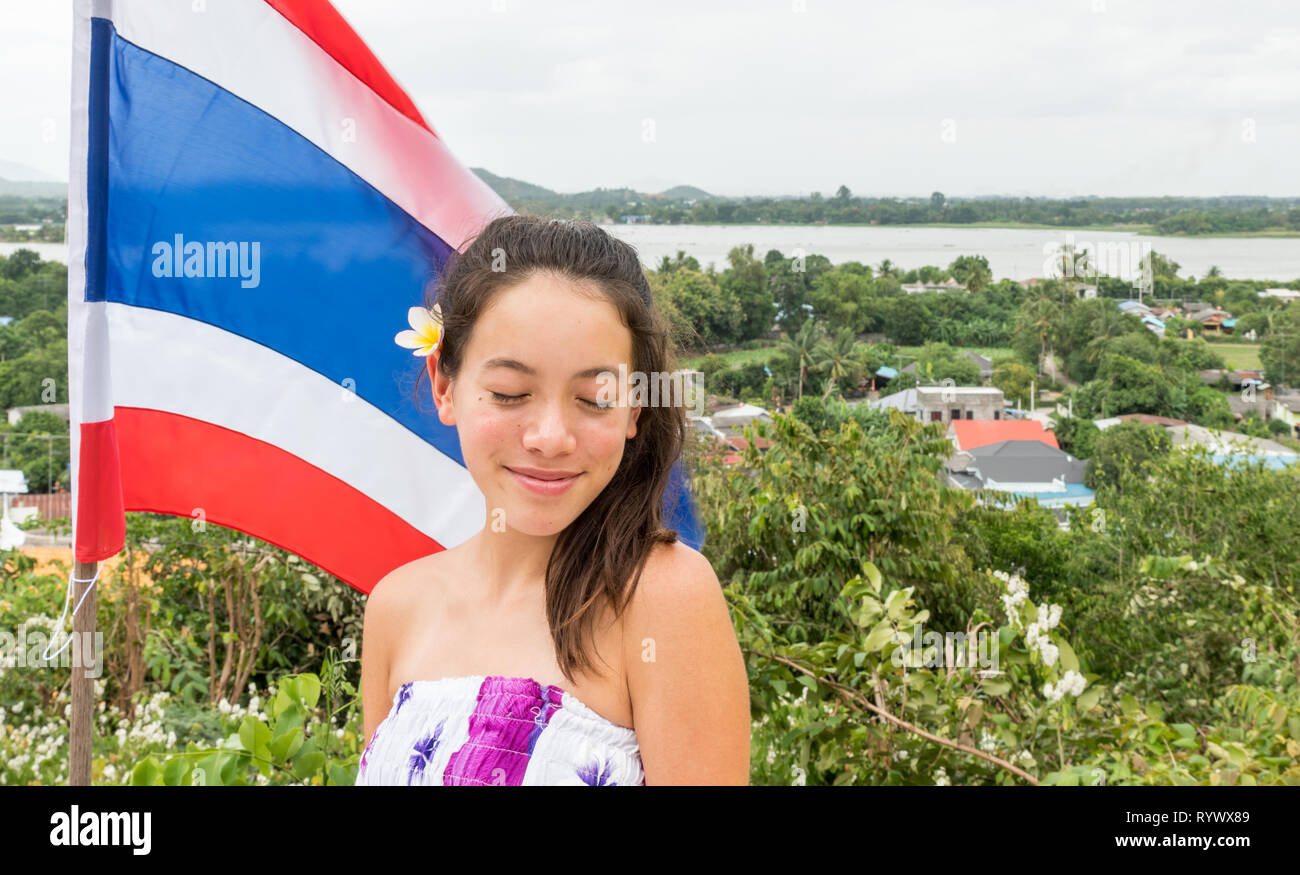 Lächelnd jugendlich mit geschlossenen Augen stand vor der Thailändischen Fahne mit der Stadt im Hintergrund Stockfoto