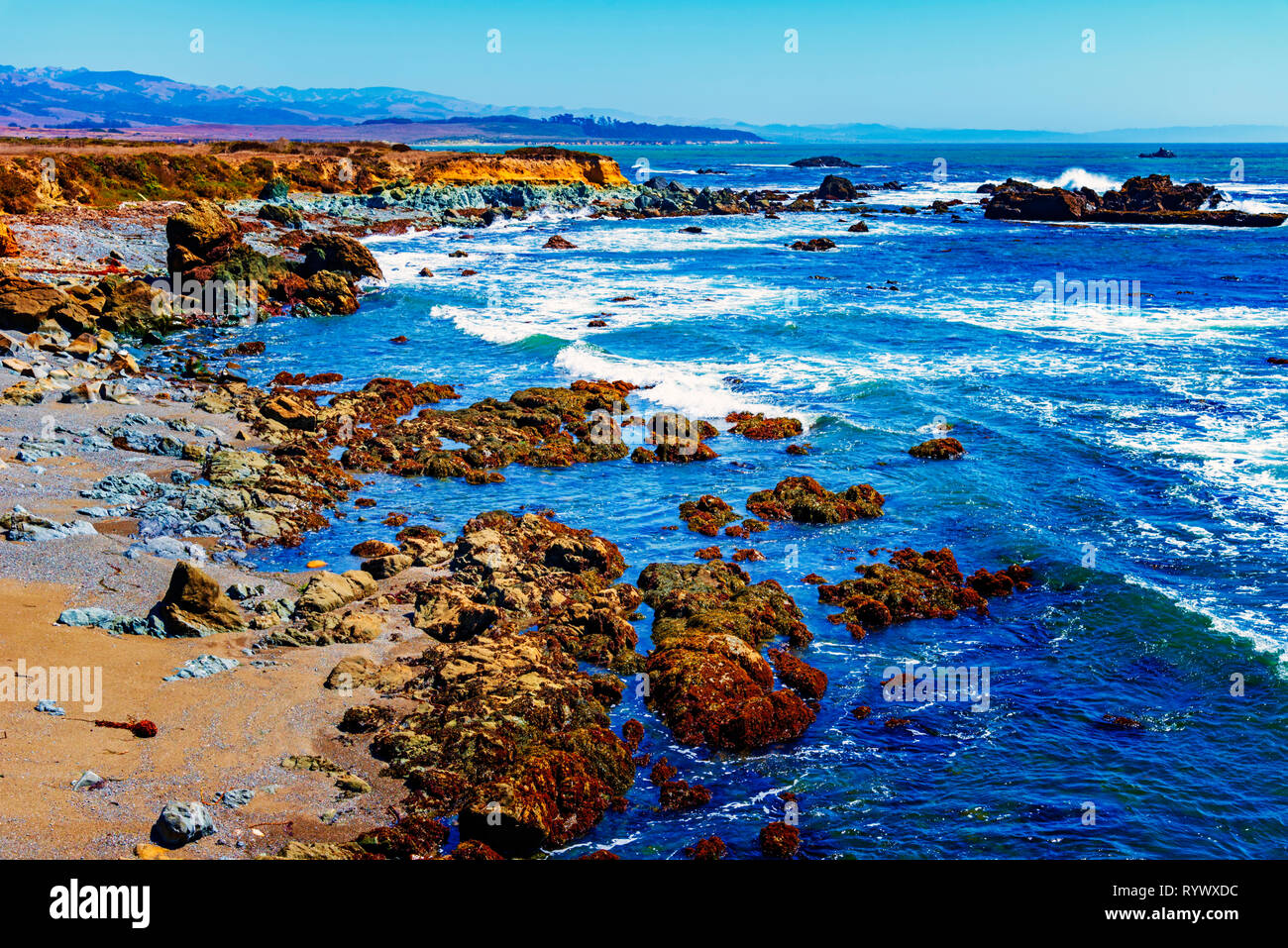 Helle, blaue Meer, Wellen, die auf der felsigen Küste mit Felsen unter strahlend blauem Himmel brechen. Stockfoto