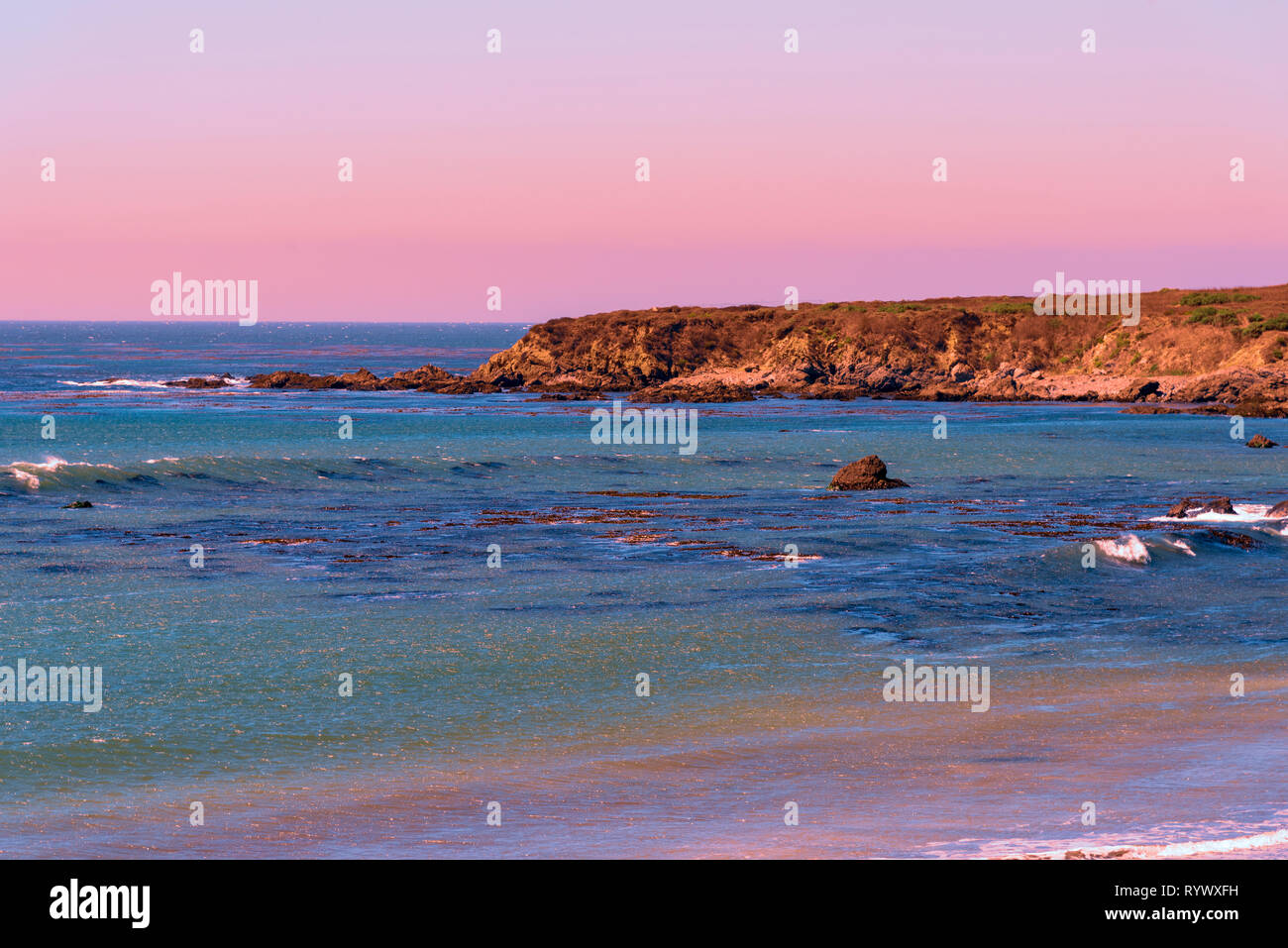 Blue Green Ocean mit kleinen Wellen, die Klippen am Meer bei Sonnenuntergang unter Blass orange, rot, pink und lila Himmel. Stockfoto