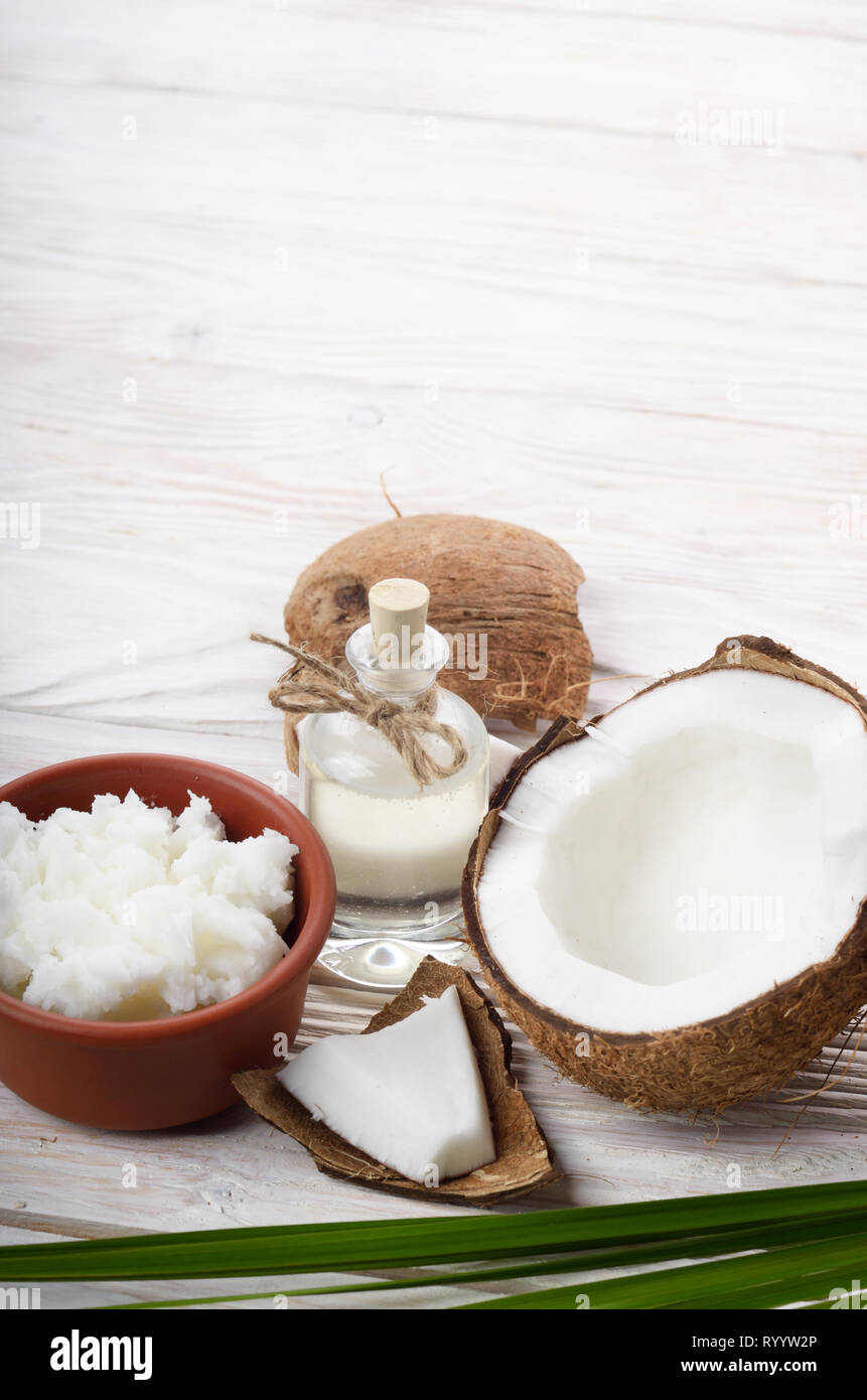 Hintergrund der Kokosnuss, Kokosnussschalen, Öl in Ton Schüssel und Glas auf weissem Holztisch Stockfoto