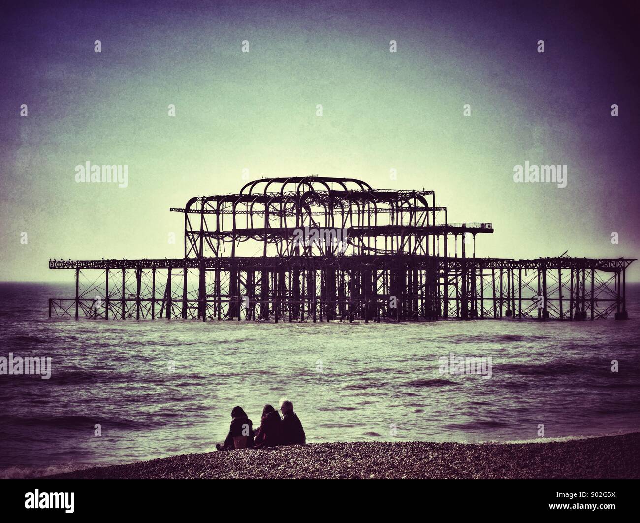 Urlauber auf Brightons Kiesstrand mit dem Untergang des West Pier im Hintergrund Stockfoto