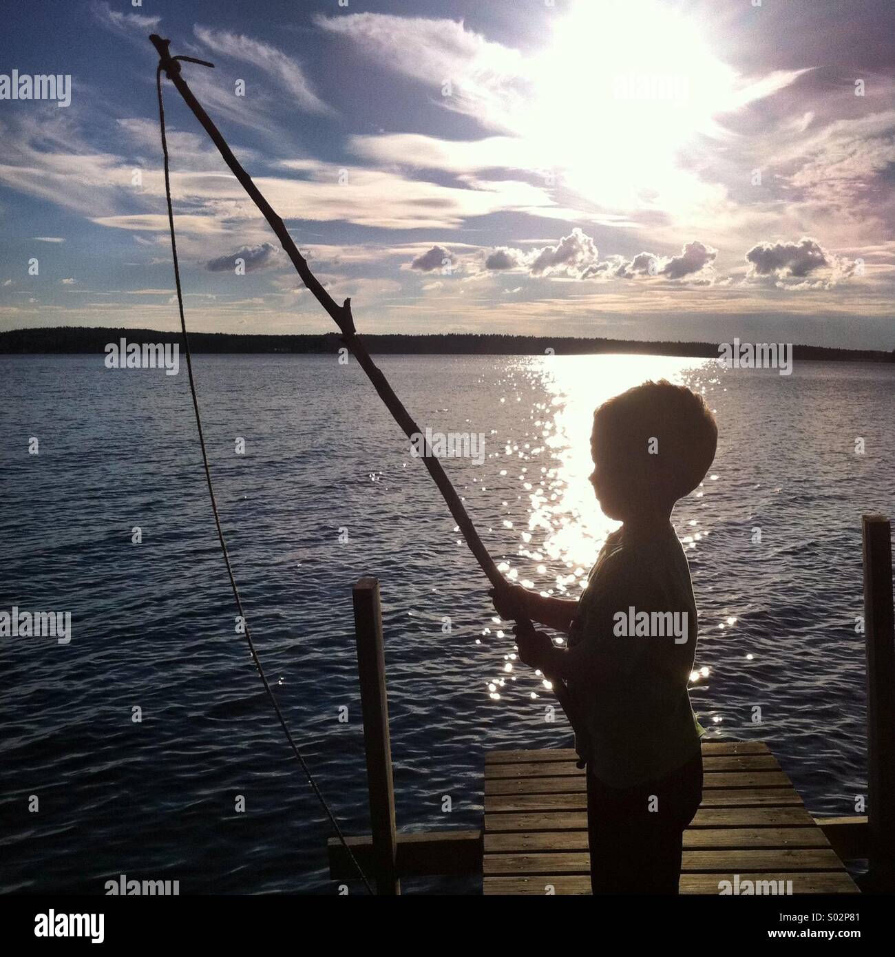 Ein kleiner Junge, der beim Angeln lernt, im Sonnenuntergang mit einer Rute aus einem Baumzweig zu fischen - Smartphone-aufgenommenes Stockfoto
