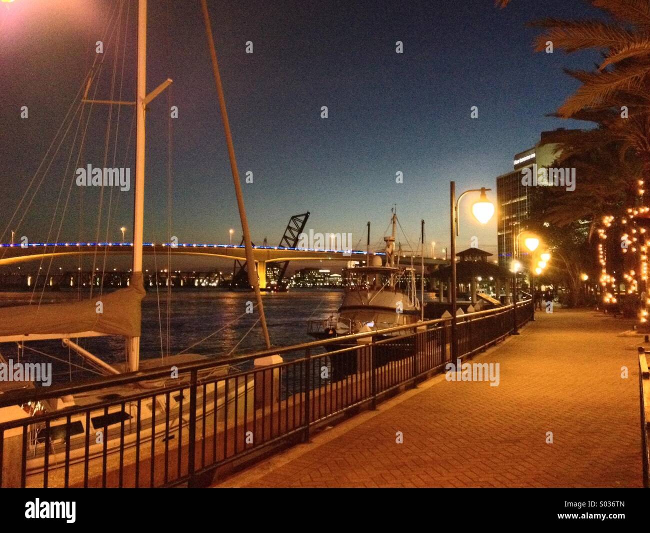 Einem frühen Abend entlang der St John am Flussufer in der Innenstadt von Jacksonville, Florida, USA, mit einem Segelboot und Brücken auf dem Wasser und nicht identifizierbare Wanderer in der Ferne Stockfoto