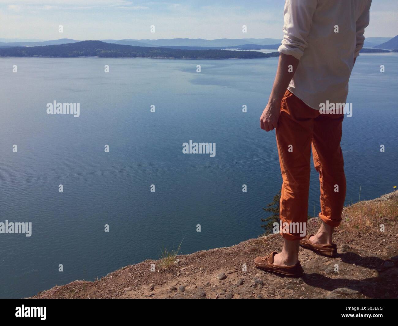 Anonyme stilvollen Mannes Oberkörper und Beinen stehend auf hohen Klippen mit Blick auf Meer und Inseln in der Sonne. Stockfoto