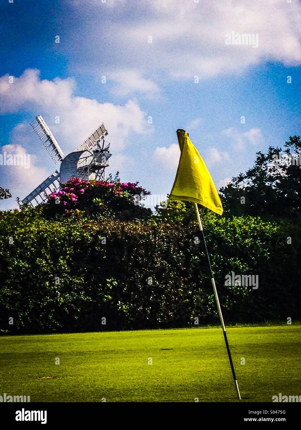 Golf-Fähnchen auf grün mit Windmühle im Hintergrund hinter Hecke Stockfoto