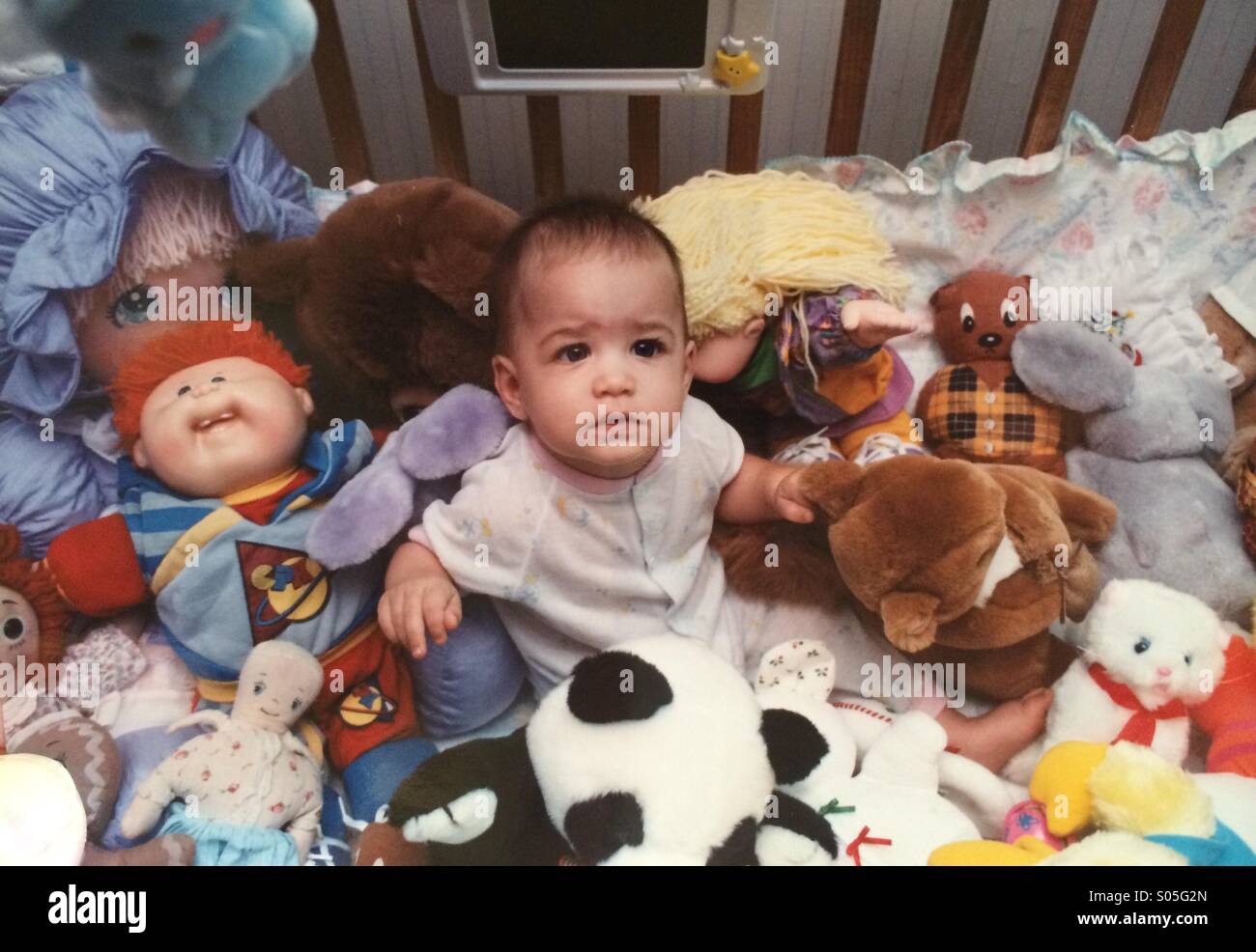 Ein Baby sitzt in einer Krippe inmitten zahlreicher ausgestopfte Tiere - Smartphone-aufgenommenes Stockfoto