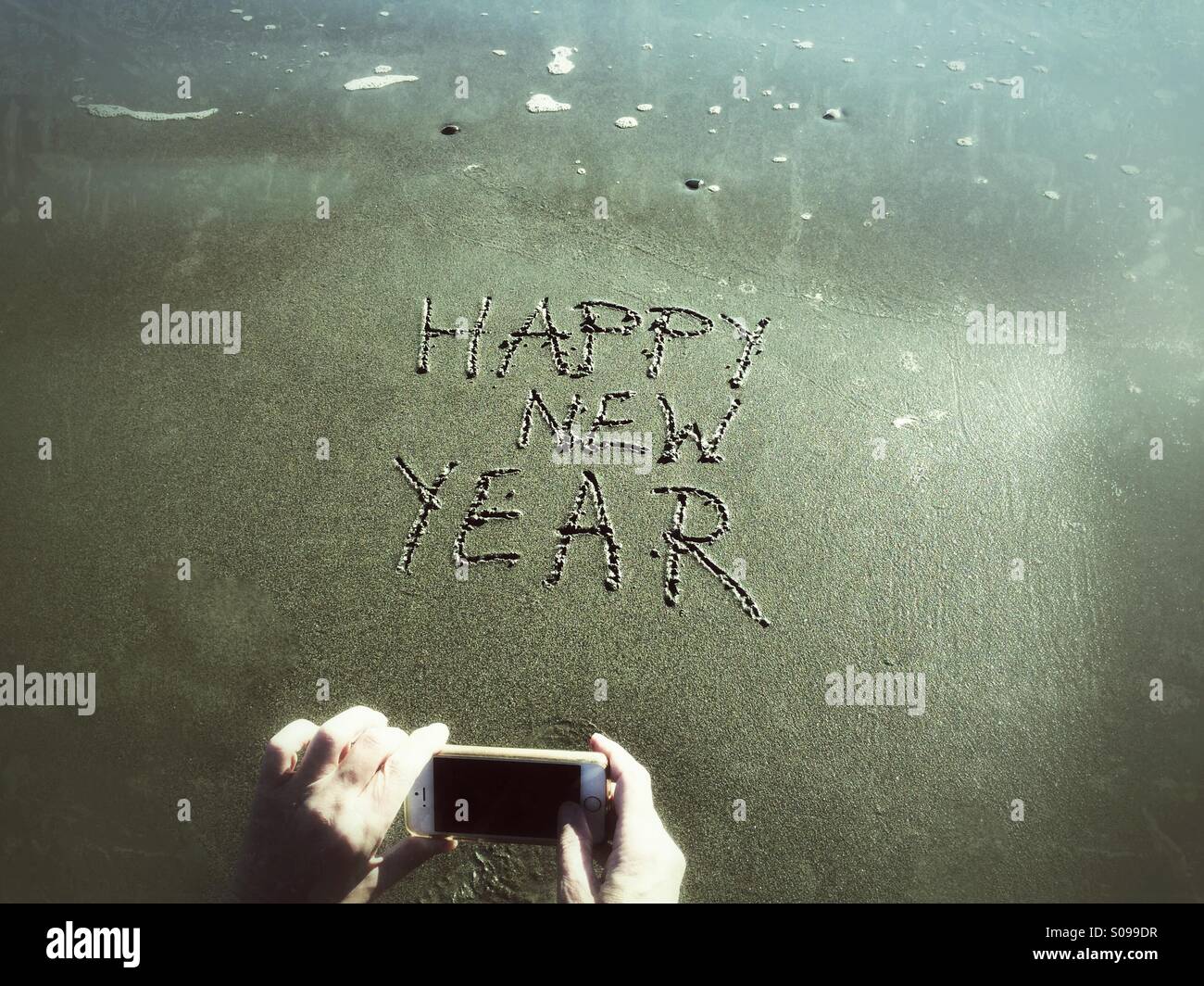 Aufnahme eines Fotos von Happy New Year, geschrieben in den Sand am Strand am Neujahrstag - Smartphone-aufgenommenes Stockfoto