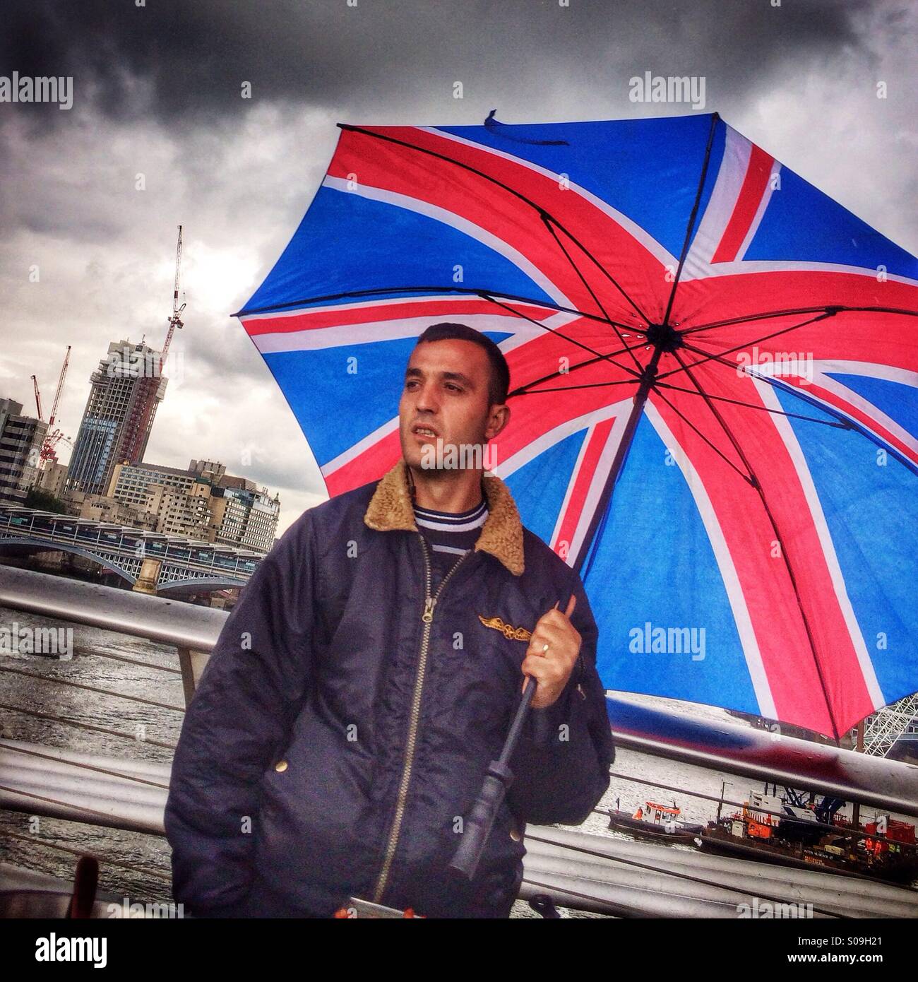 Ein Mann trägt einen bunten Regenschirm mit einem union Jack-Design hebt sich von London Gewitterhimmel. Stockfoto
