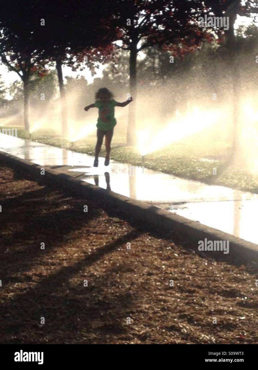 Kleines Mädchen mit Sprinkler hinter ihr springen Stockfoto