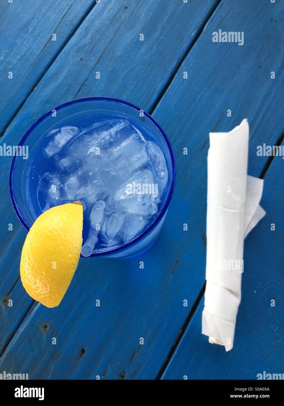 Longdrinkglas mit Eiswasser mit Zitrone und Stroh und eine Serviette, auf einem blauen Holztisch Stockfoto