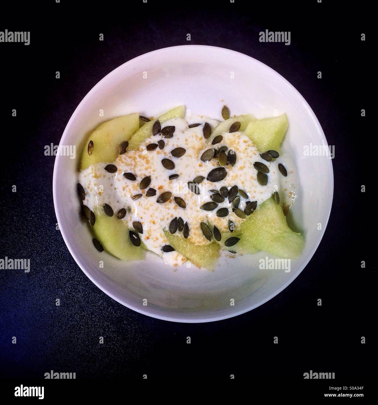 Galia Melone mit Joghurt, braunem Zucker und Kürbiskerne in einer weißen Schüssel auf einem schwarzen Tisch. Stockfoto