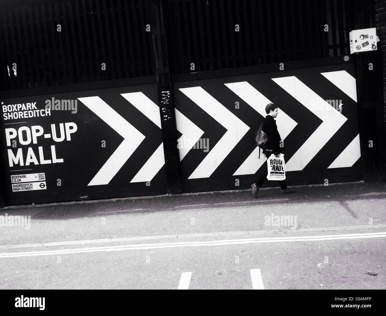 Ein Mann gekleidet in schwarz mit einer weißen Rough Trade Plattenladen Tasche vorbei an einem großen schwarzen und weißen Chevron Schild führt zu Boxpark Pop geht Up Mall in der Nähe von Brick Lane in Shoreditch, London, England. Stockfoto