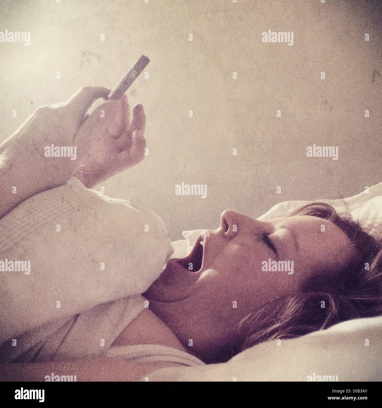 Eine gähnende Frau im Bett mit einem Mobiltelefon im Bett liegend Stockfoto