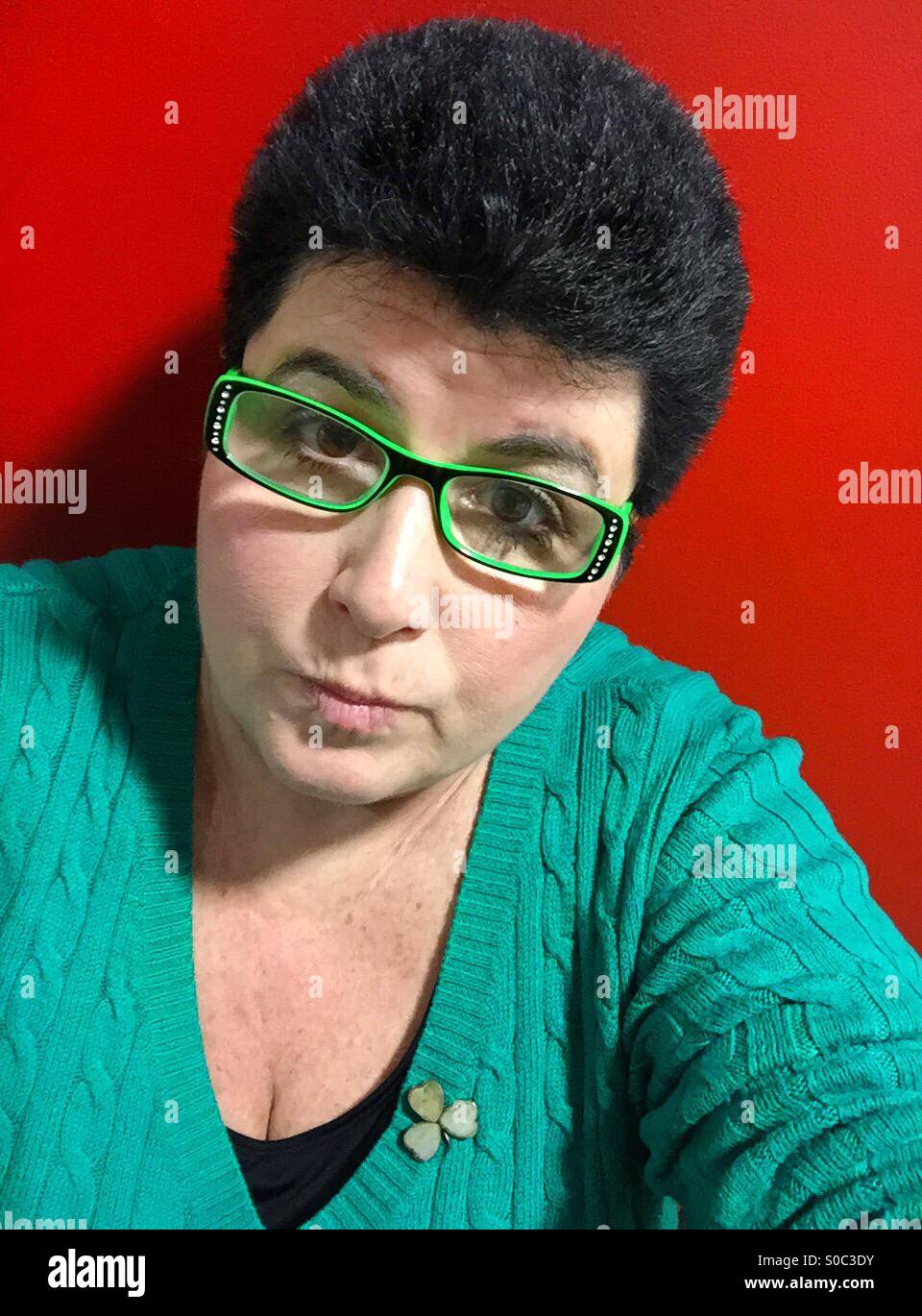Eine Frau mit schwarzen Haaren tragen grün und ein Kleeblatt Pin, mit Neon-grün-Brille, gegen eine helle rote Wand. - Smartphone-aufgenommenes Stockfoto