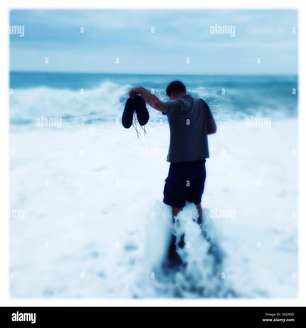 Silhouette der Mann, der in Wellen auf einem dunklen Sandstrand hält Schuhe Stockfoto