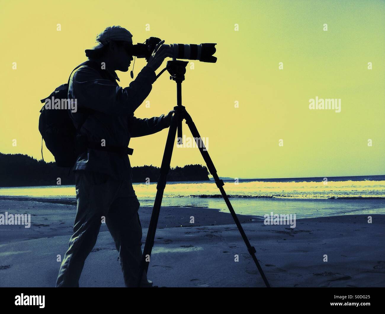 Mann mit Kamera auf Stativ an einem Strand - Smartphone-aufgenommenes Stockfoto