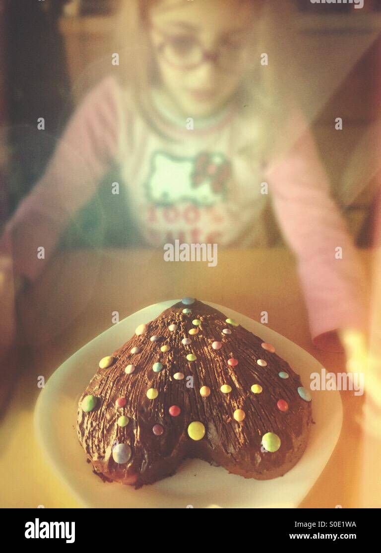 Kleine blonde Mädchen Blick auf ihr Hart geformte Schokolade Kuchen mit Smarties. Herzlichen Glückwunsch zum Geburtstag. Stockfoto