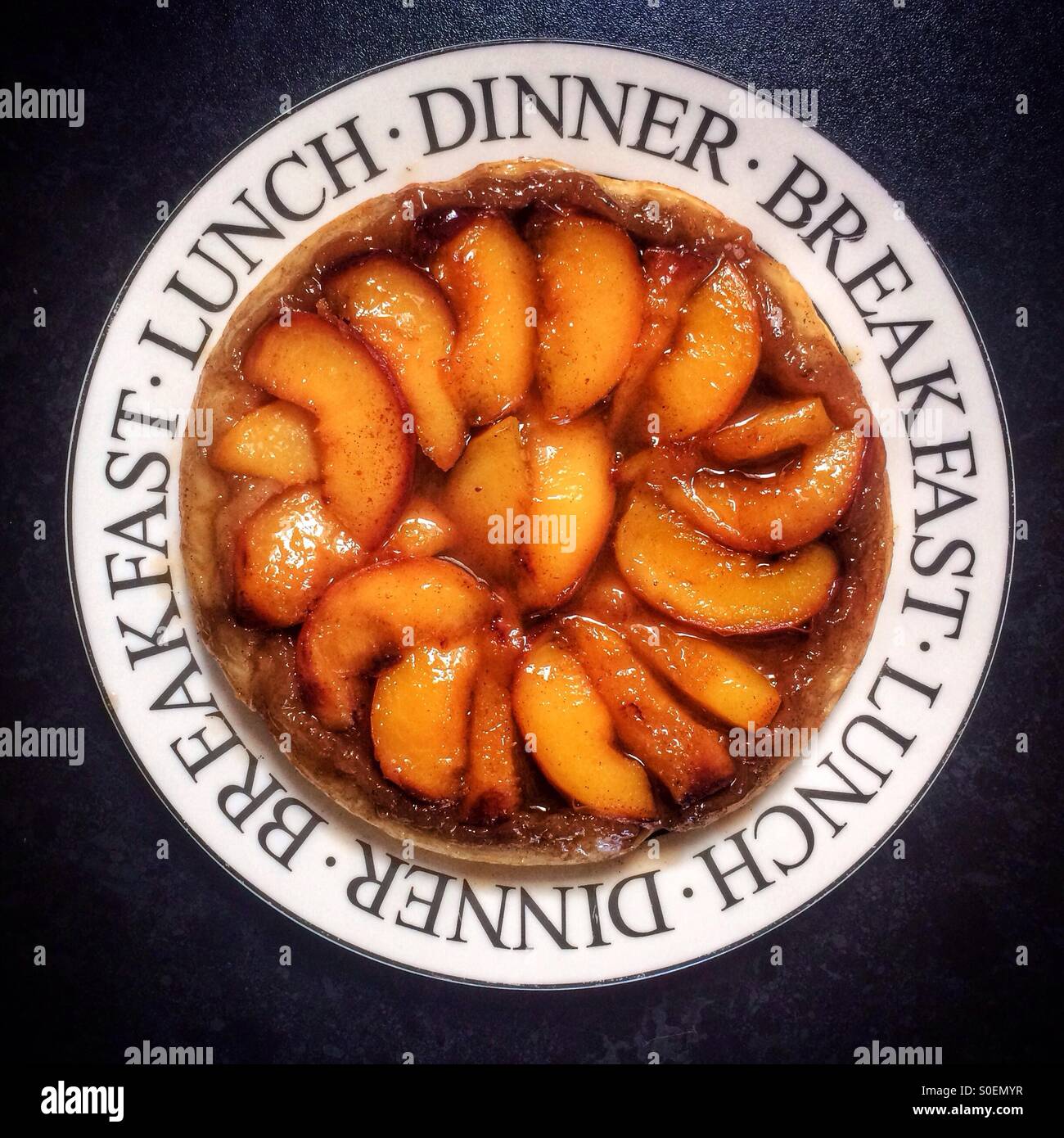 Pfirsich-Tarte Tatin auf einem weißen Teller dekoriert mit den Worten Frühstück, Mittag- und Abendessen gegen einen schwarzen Hintergrund. Stockfoto