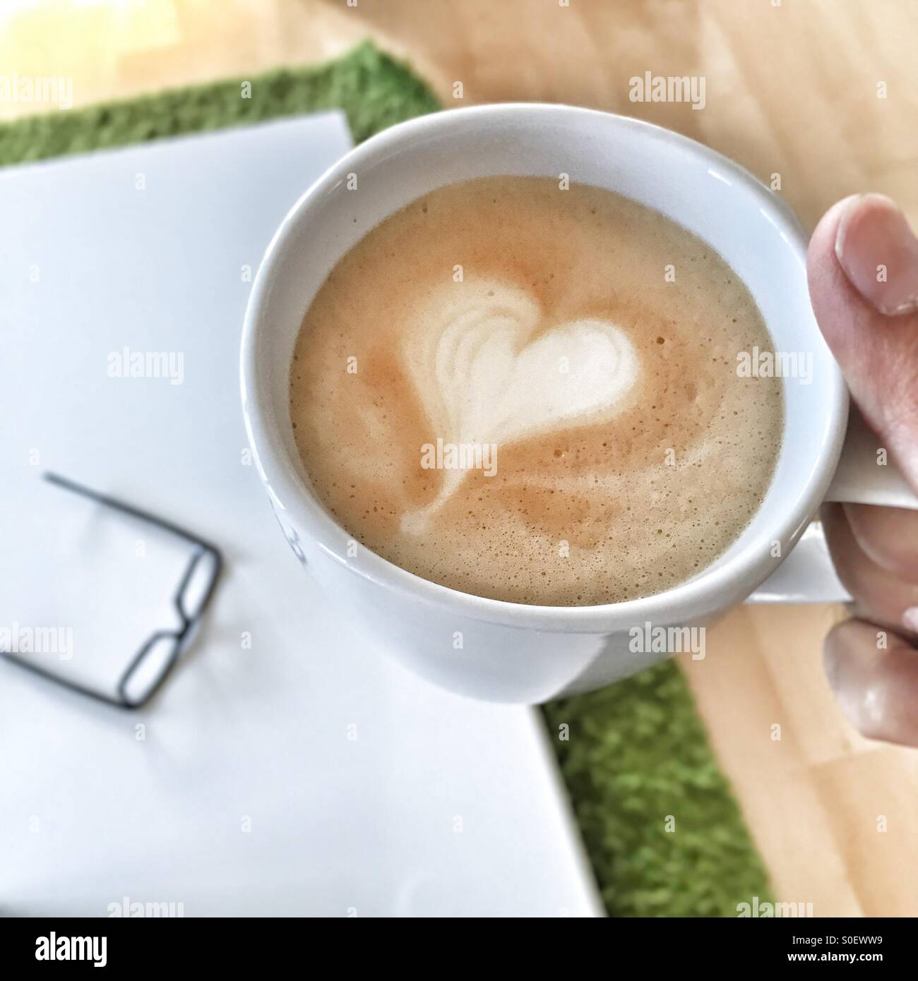 Hand hält eine Tasse Kaffee, Brille auf dem Tisch im Hintergrund. - Smartphone-aufgenommenes Stockfoto