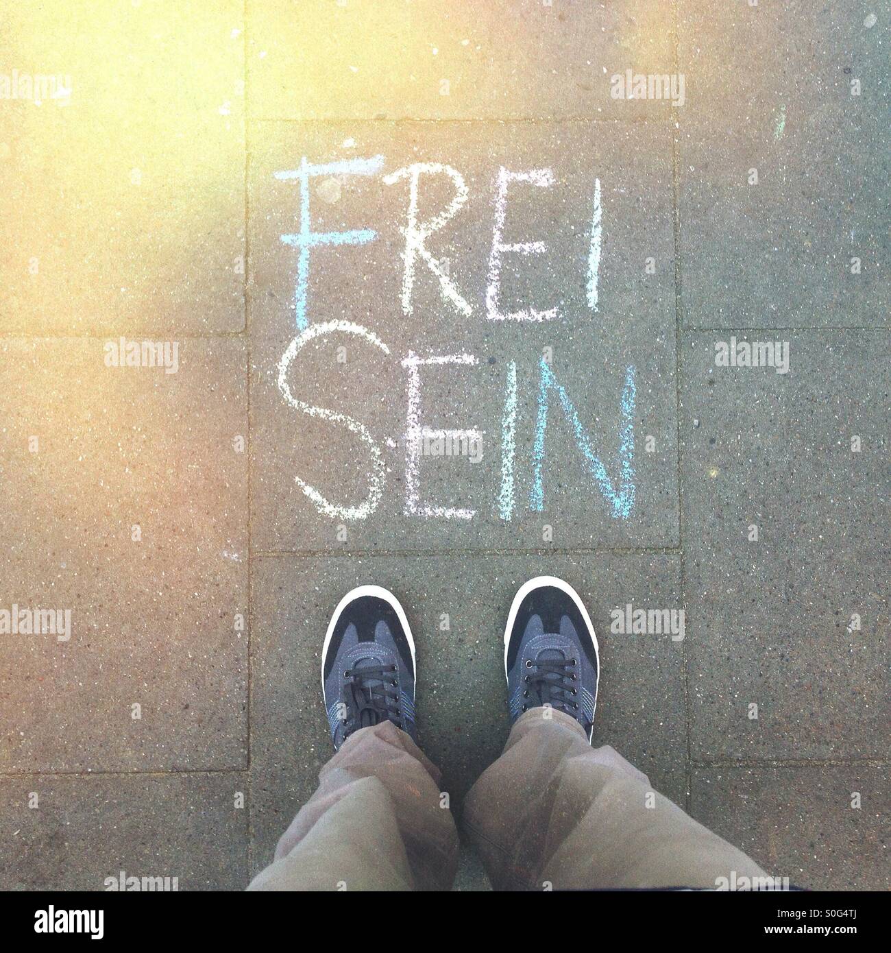 A mans Füße neben die Worte "Frei Sein" (Be Free) auf dem Bürgersteig in deutscher Sprache mit bunter Kreide geschrieben steht Stockfoto