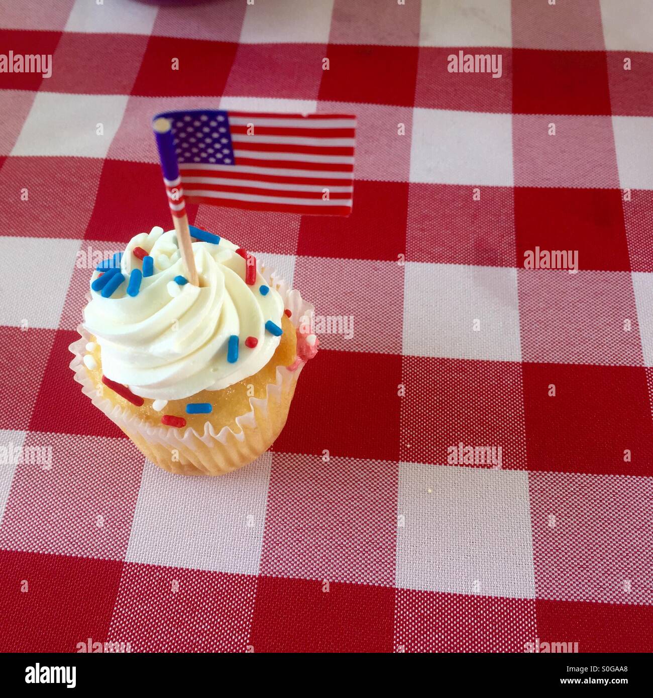 Ein niedlicher weißen Cupcake bestreut mit roten, weißen und blauen Streusel mit einer amerikanischen Flagge sitzt auf einem karierten Tischdecke. Stockfoto
