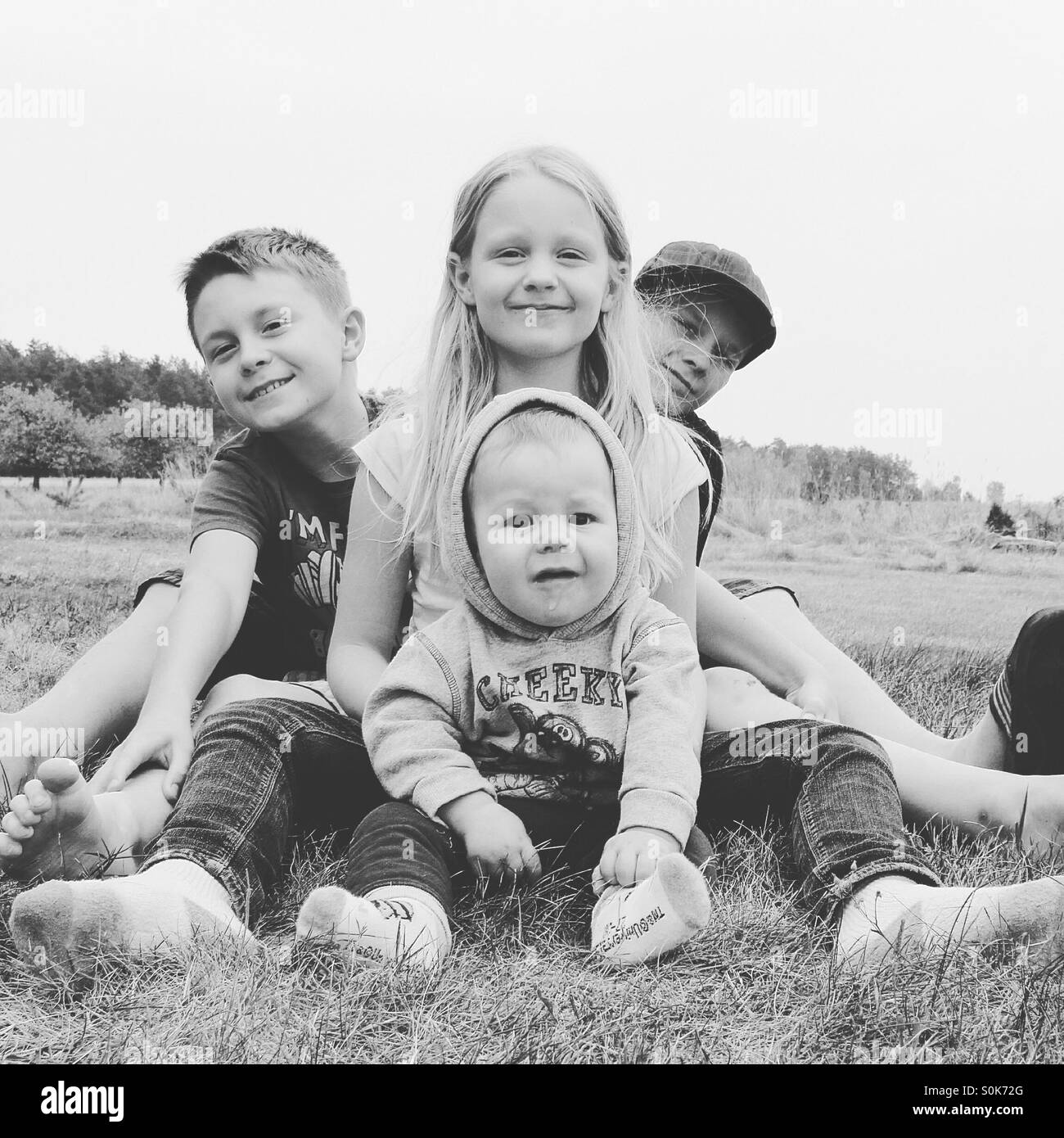 Vier Geschwister Alter 8 Monate bis 12 Jahre sind posieren und Lächeln für ein Familienfoto, draußen, sitzen auf einer Wiese Stockfoto