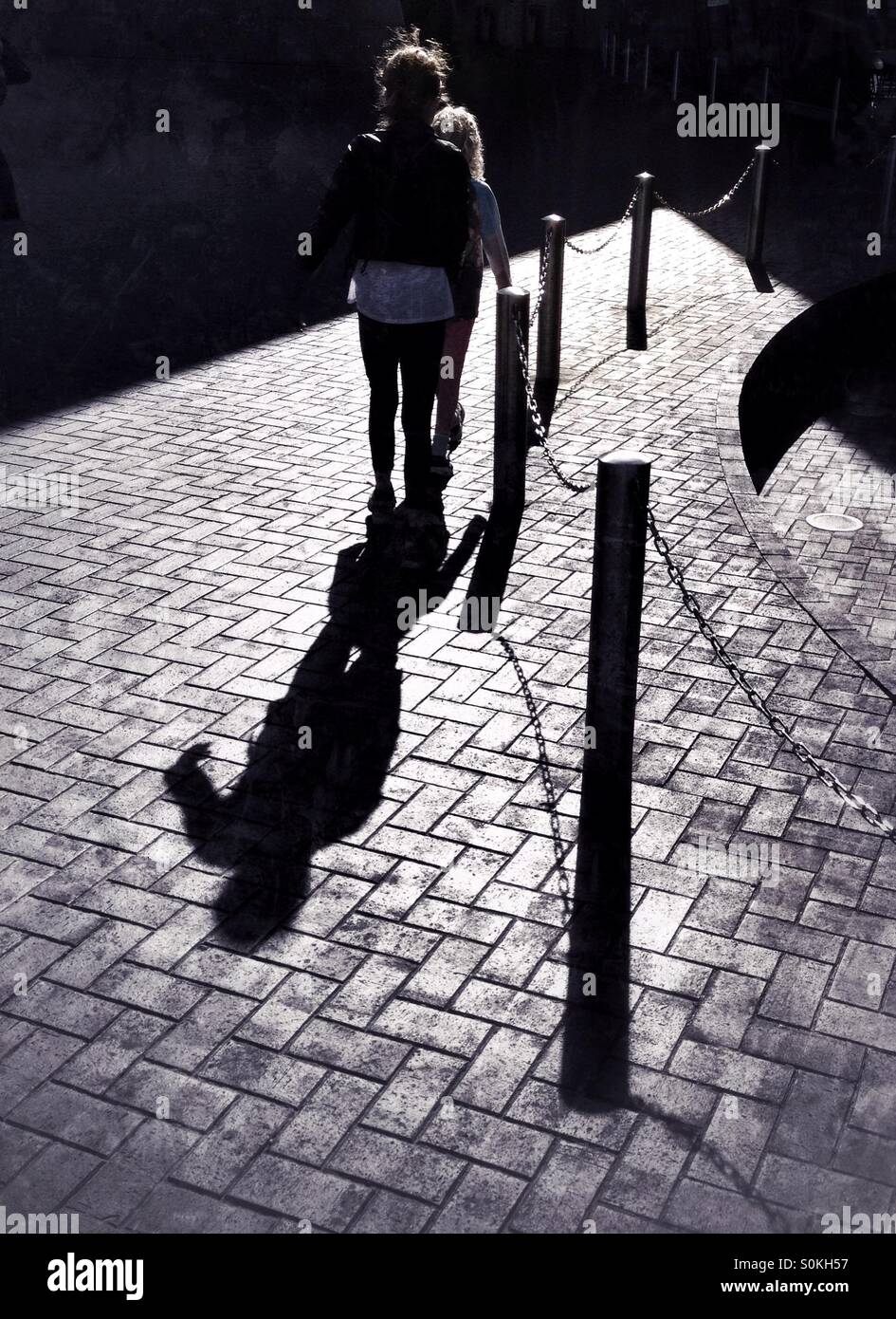 Zwei junge Kinder werfen Schatten auf gepflasterten Weg durch Poller Stockfoto