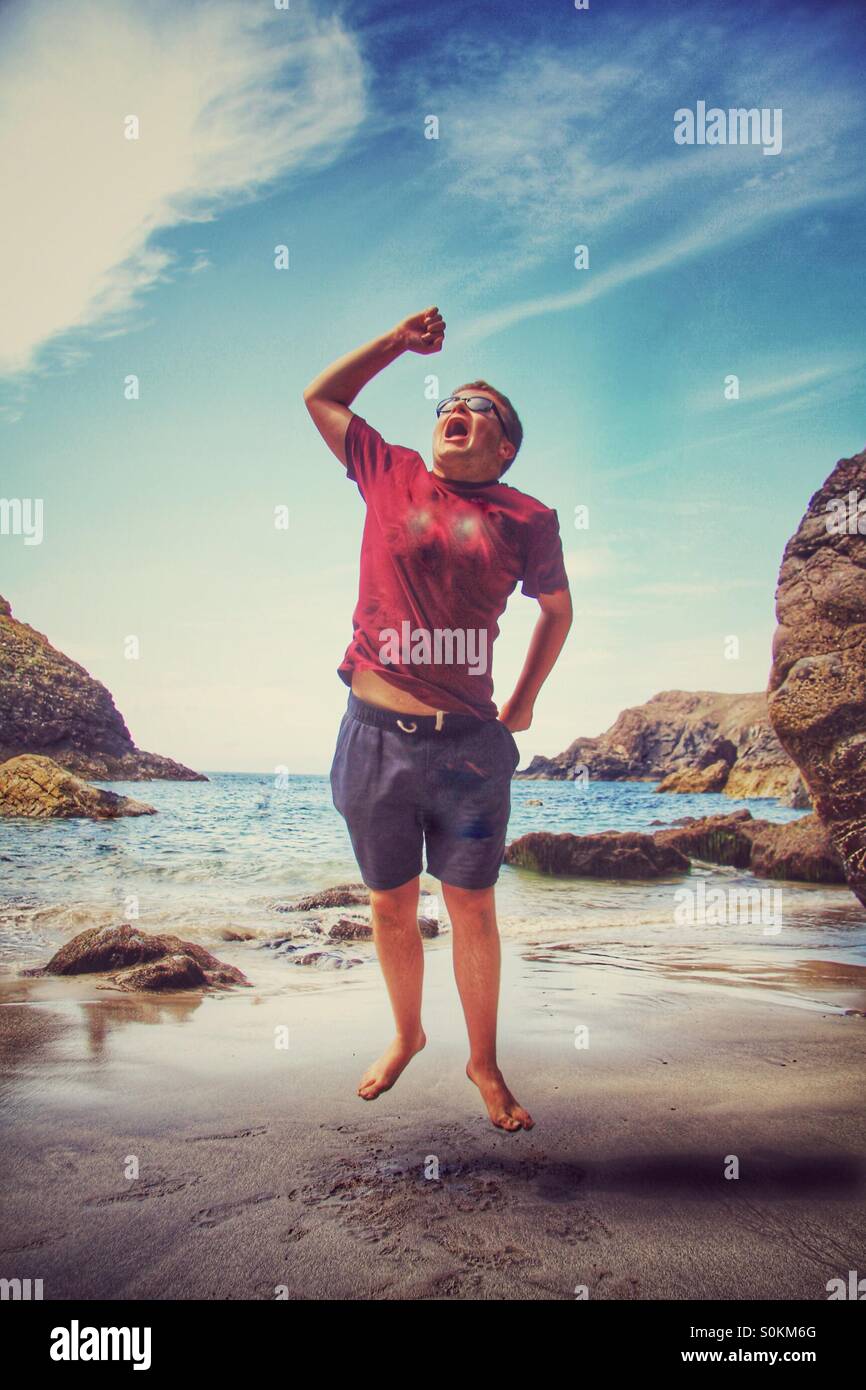 Ein Junge auf einem sandigen Strand springen vor Freude. Ein Cornish Strand mit dem Meer hinter ihm. Stockfoto