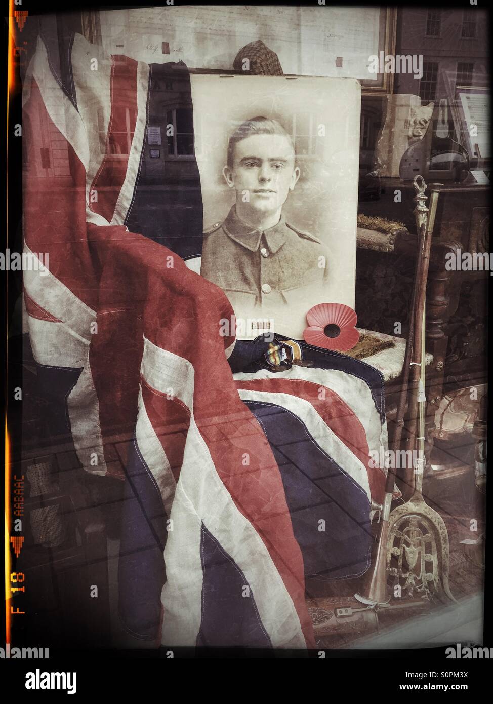 Ein Schaufenster, Armistance Day, 11. November gedenken. Der Union Jack Flagge wird über einen Stuhl und neben einem roten Mohn, Krieg Medaillen und ein fotografisches Porträt eines jungen Soldaten drapiert. - Smartphone-aufgenommenes Stockfoto