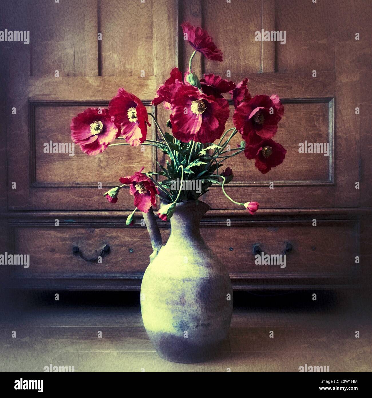 Rote Mohnblumen in einer rustikalen Keramikvase vor antiker Holzkulisse angeordnet, stilisiertes Stillleben mit zeitloser, nostalgischer Stimmung. Stockfoto