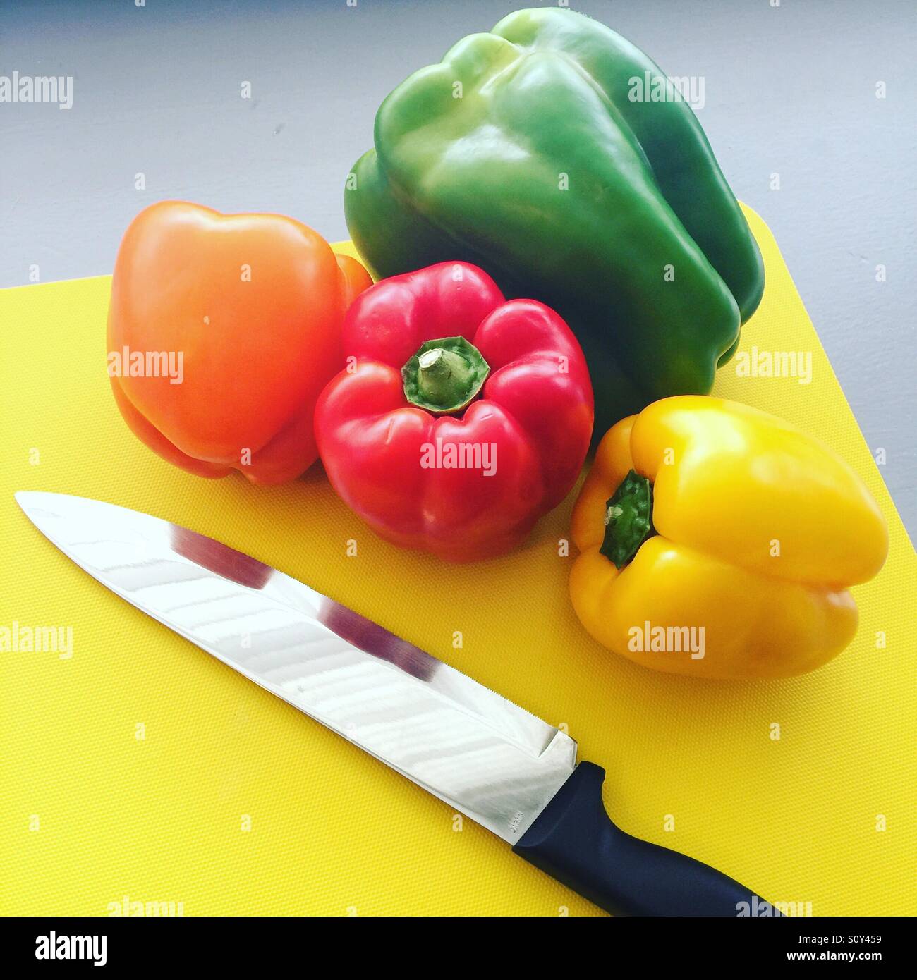 Eine rote, orangefarbene, gelbe und grüne Paprika mit einem Messer auf einen gelben Hackstock. - Smartphone-aufgenommenes Stockfoto