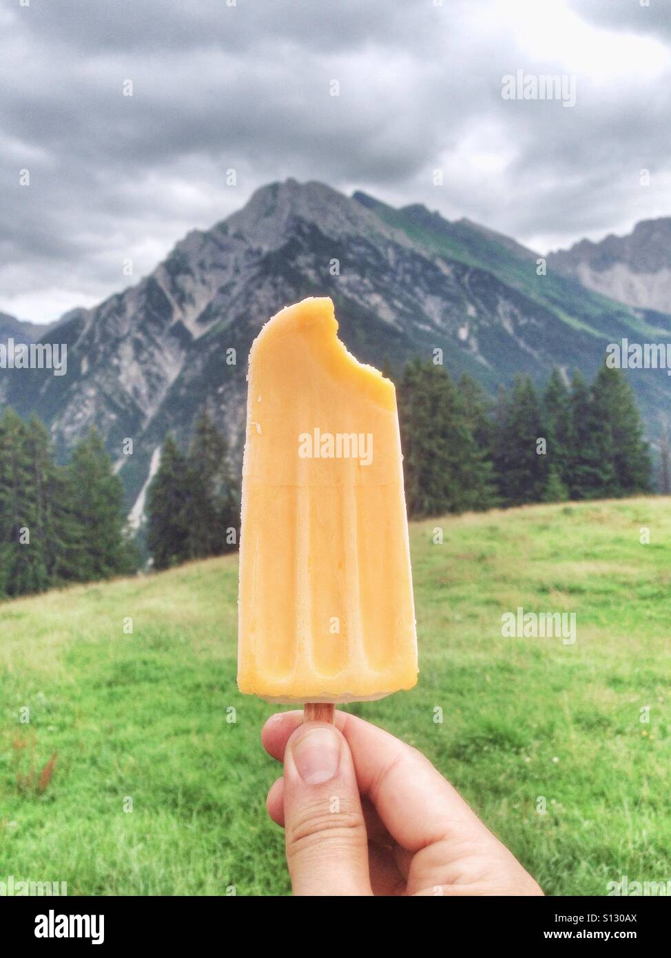 Eis am Stiel in einer männlichen kaukasischen Hand vor einem Berg und einer grünen Wiese in den österreichischen Alpen - Smartphone-aufgenommenes Stockfoto