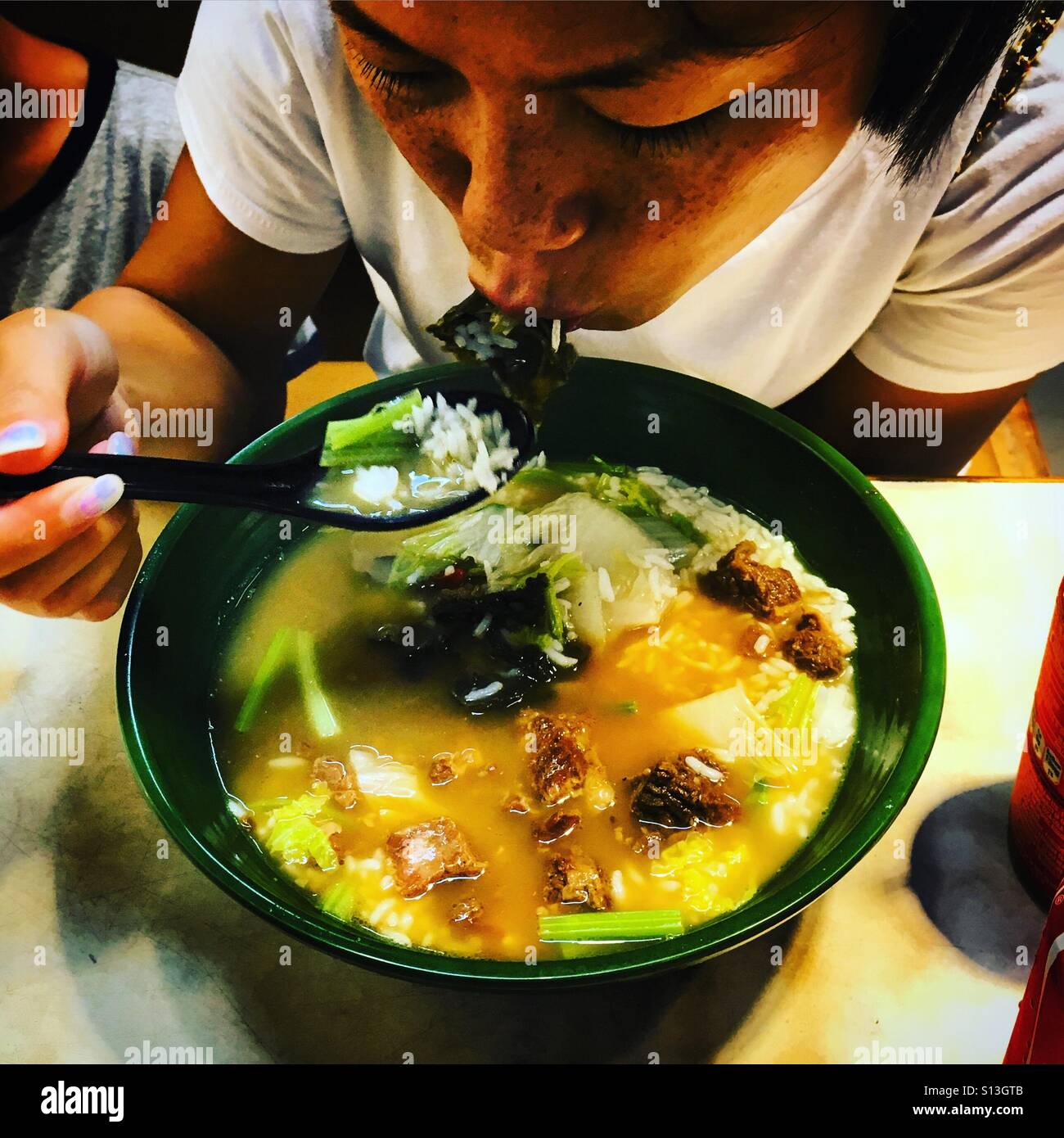 Asiatisch-chinesische Frau isst heiße Nudelsuppe in einer Schüssel, während sie in ihren Löffel pustet, um sie abzukühlen - Smartphone-aufgenommenes Stockfoto