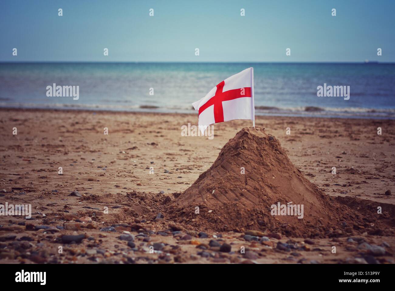 Eine große Sandburg am Sandstrand mit einer England-Flagge weht im Wind und das Meer im Hintergrund. - Smartphone-aufgenommenes Stockfoto