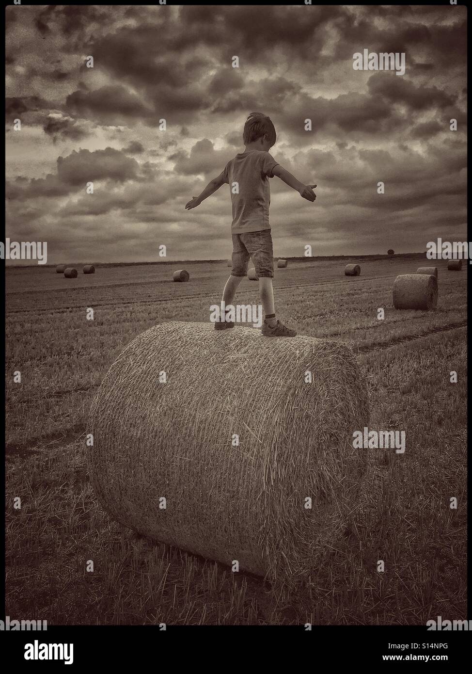 Ein 7 Jahre alter Junge drückt einen Heuballen zwischen seinen Beinen, er balanciert auf einem anderen Heuballen! Die Einstellung ist ein stürmischer Feld im Süden Englands. Ein Foto, um die Freuden der Kindheit zu veranschaulichen - Smartphone-aufgenommenes Stockfoto