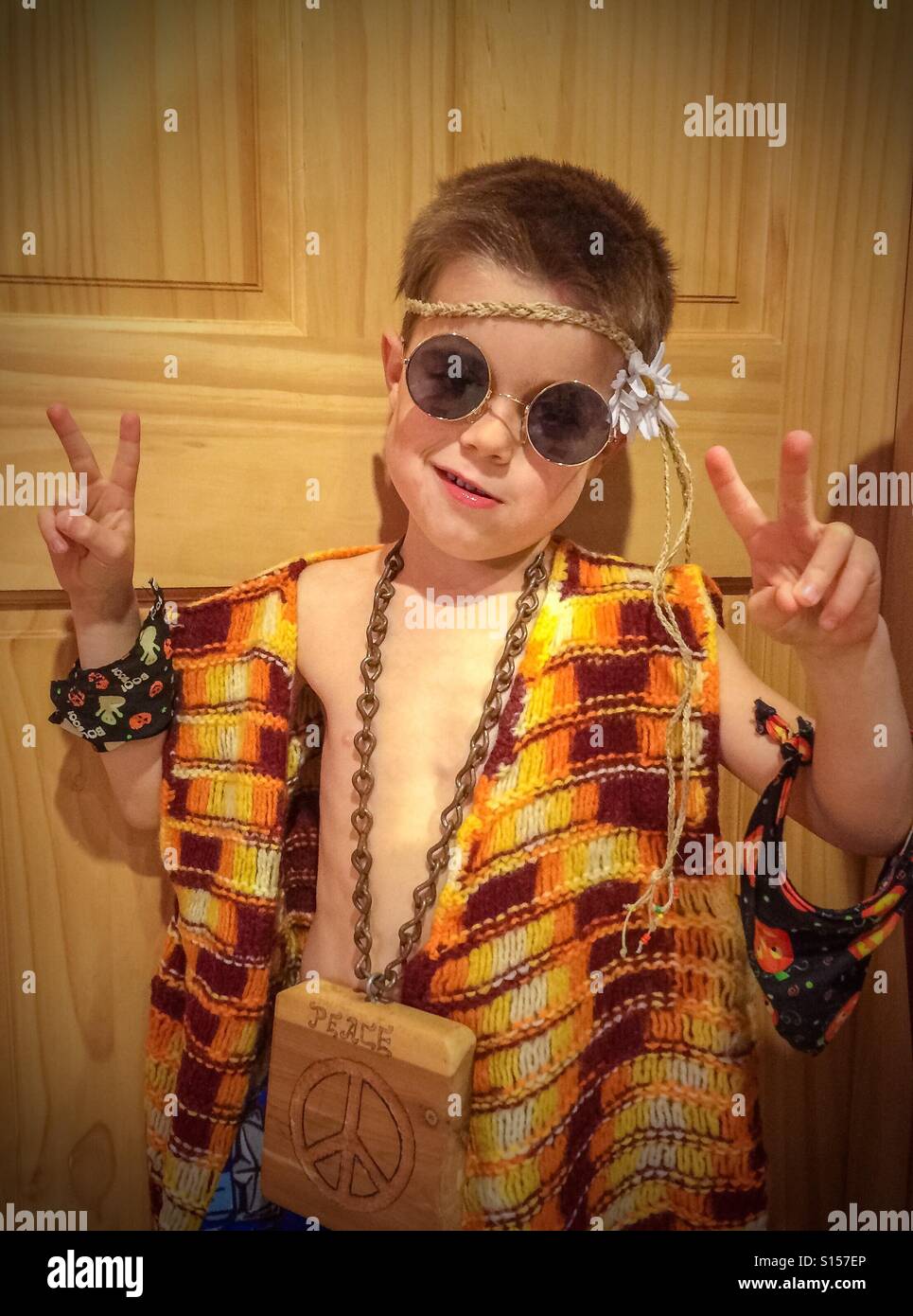 Ein 6-jähriger Junge kleidet sich in Stücke eines Hippie-Kostüms von einem vergangenen Halloween. Er macht das Friedenszeichen mit beiden Händen. - Smartphone-aufgenommenes Stockfoto