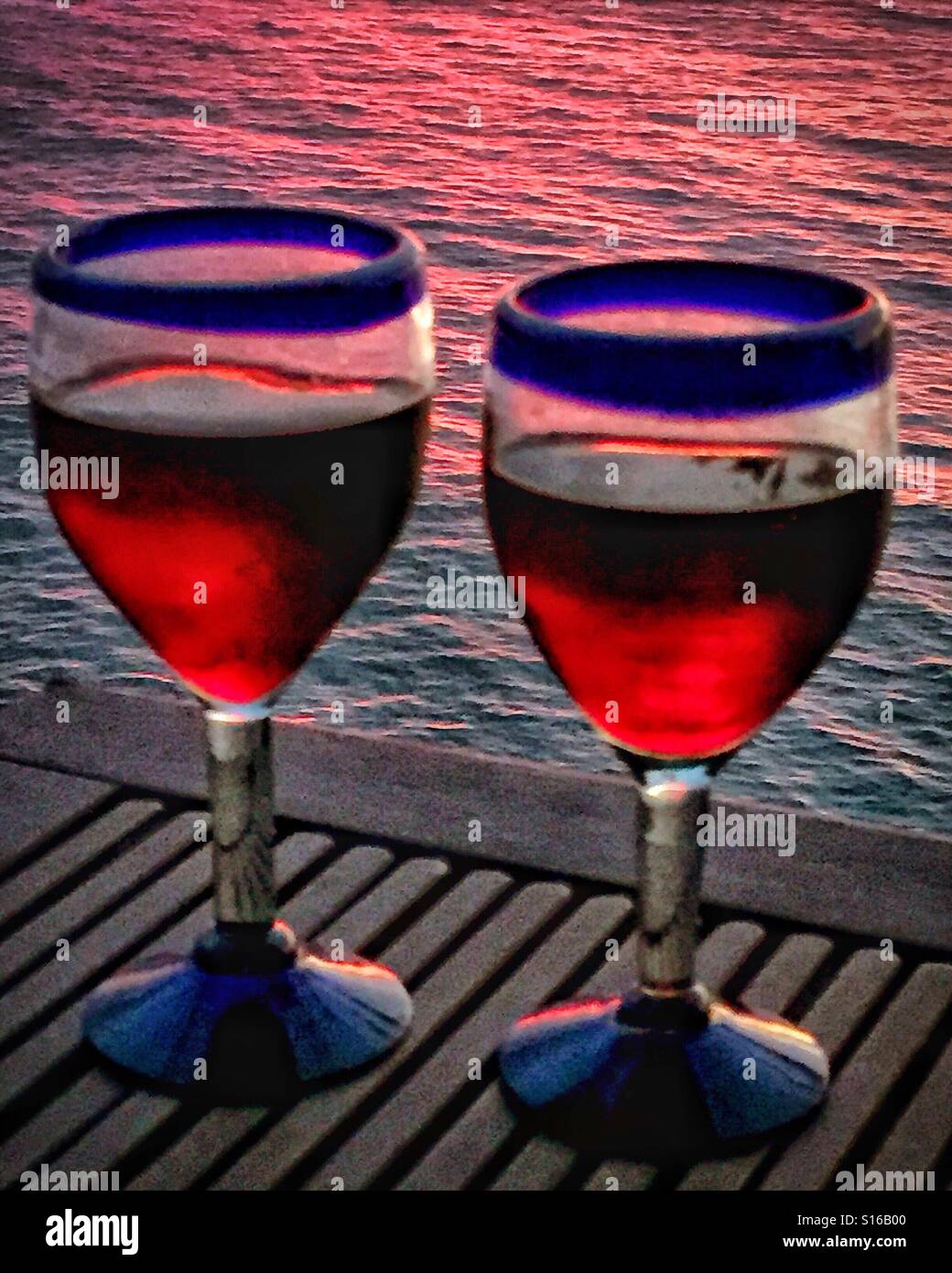Zwei authentische mexikanische blau umrandeten Hand geblasene Gläser mit Rotwein gefüllt sind und liegen auf einem Tisch aus Teakholz mit Blick auf den Pazifischen Ozean bei Sonnenuntergang. Stockfoto