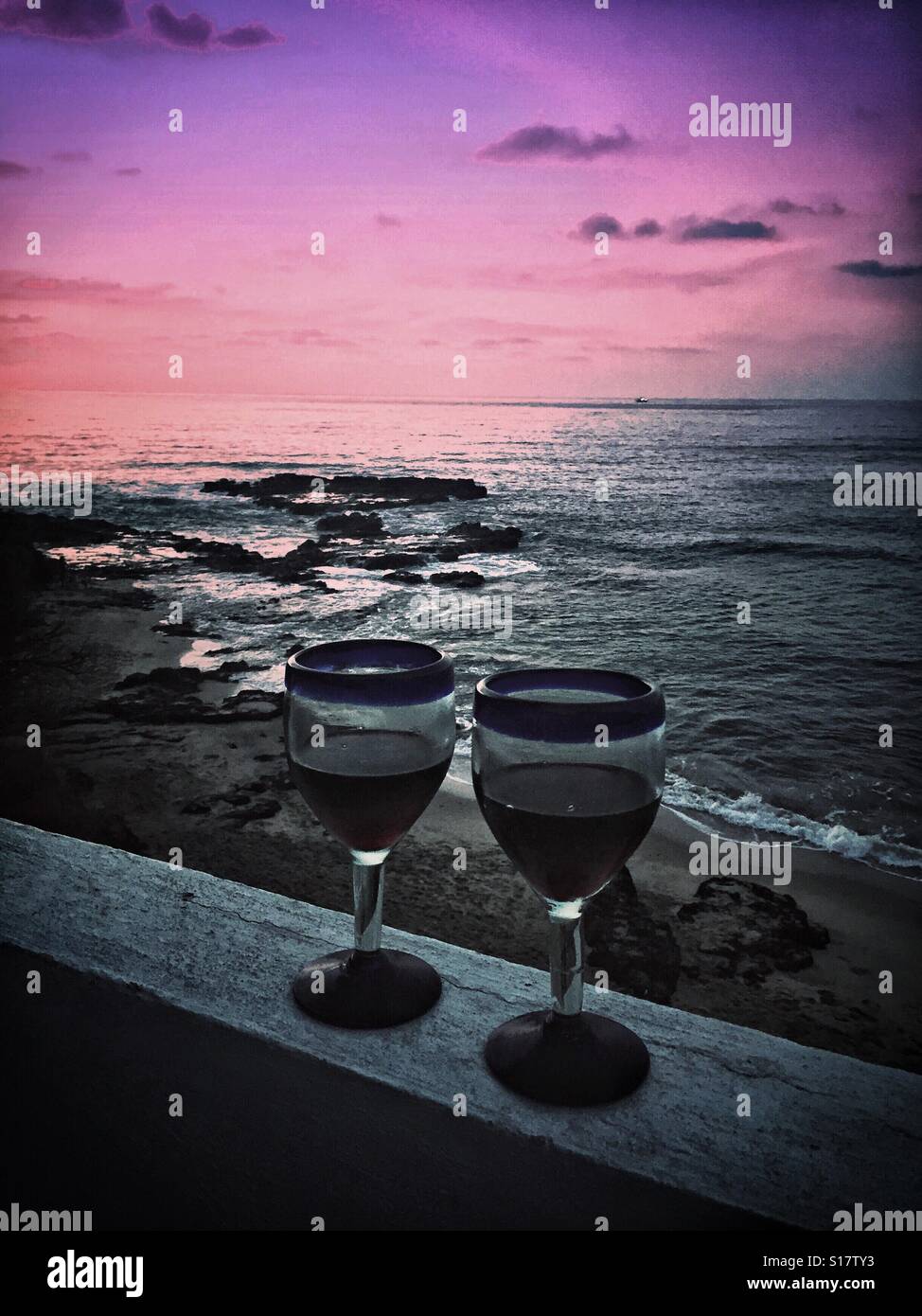 Zwei Gläser Rotwein im mexikanischen mundgeblasenen Weingläsern serviert auf einem Balkon mit Blick auf die zerklüftete Küste von Nayarit bei Sonnenuntergang. Stockfoto