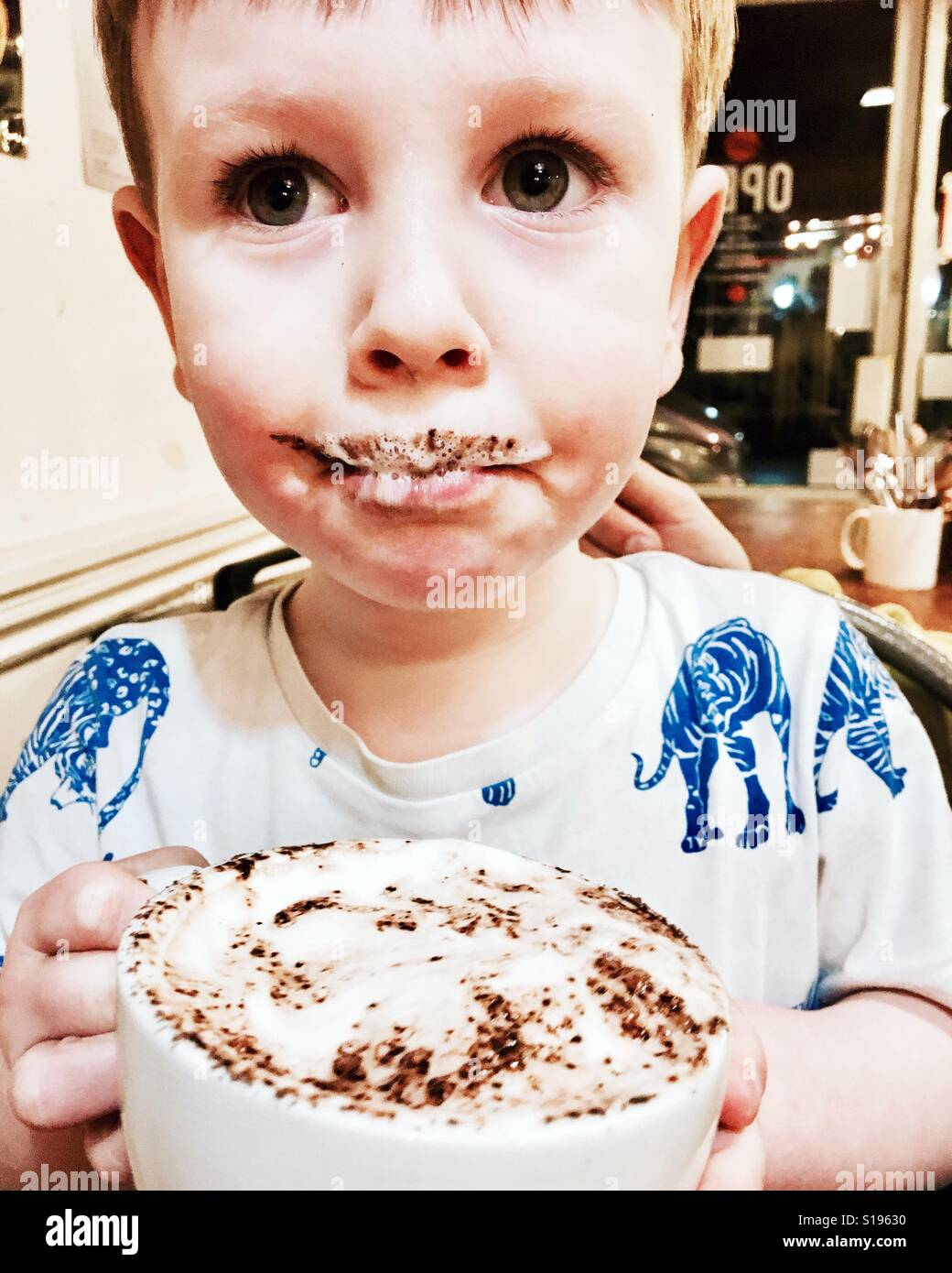Ein Kleinkind junge trinkt aus einem Babycino und bekommt einen milchigen schokoladigen Schnurrbart Stockfoto