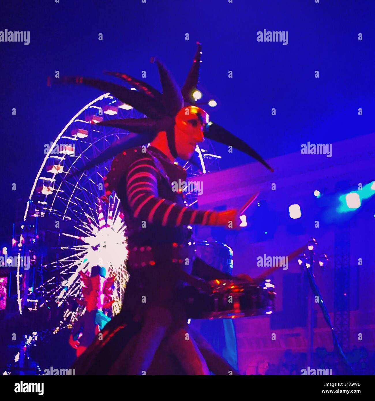 Schlagzeuger im Jester Kostüm führt für eine Nacht Menschenmenge vor einem Riesenrad an der schönen Carnaval. Stockfoto