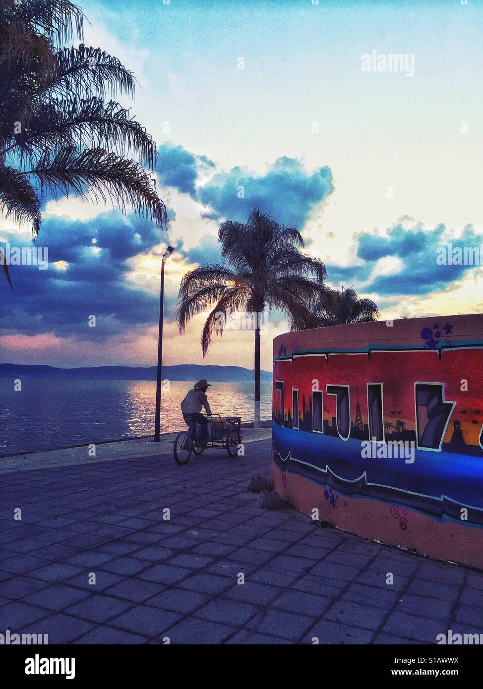 Ein Verkäufer auf einem Fahrrad fährt mit dem Ajijic Malecon bei Sonnenuntergang. Stockfoto