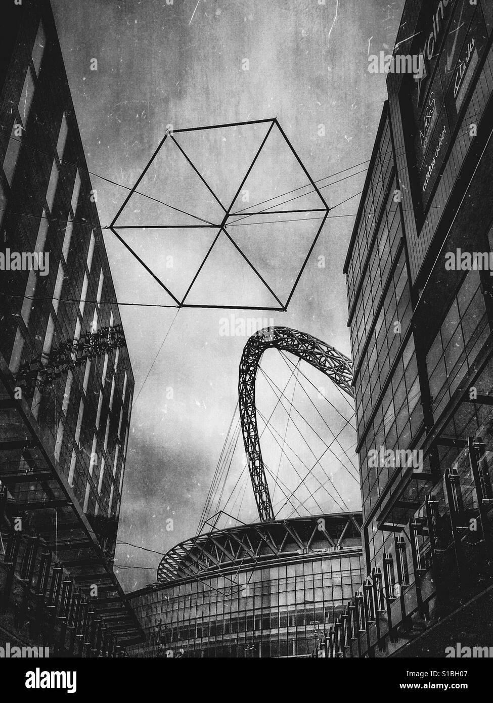 Schwarz / weiß Foto einer geometrischen Würfels Skulptur vor Wembley Stadion gesehen zwischen den Gebäuden der Londoner Designer Outlet, London, England, UK. Stockfoto