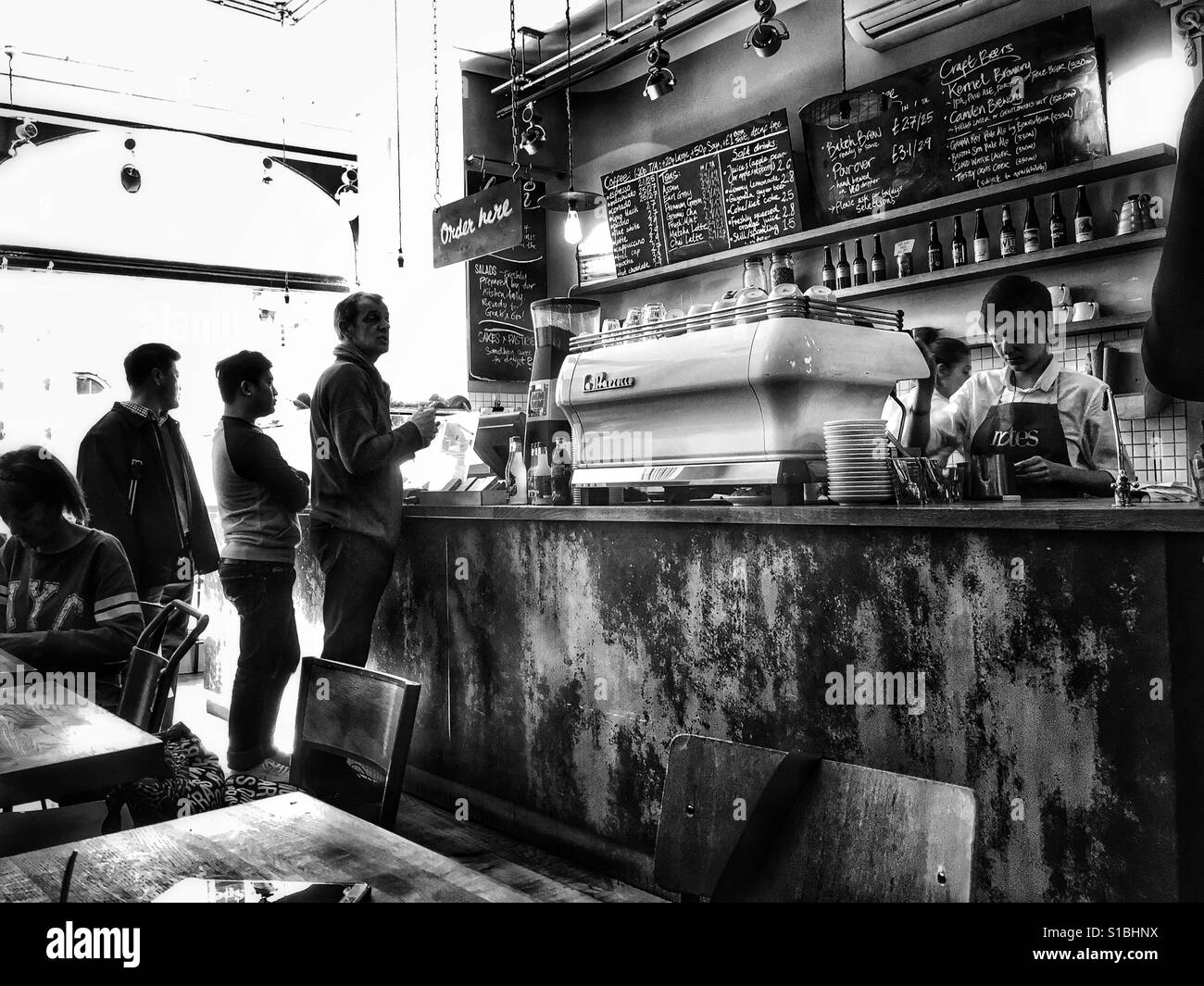 Schwarz / weiß Foto des Kunden Warteschlangen für den Kaffee von der Espresso-Maschine als die Barista macht ihre Getränke auf Notizen Kaffee, London, England, UK. Stockfoto